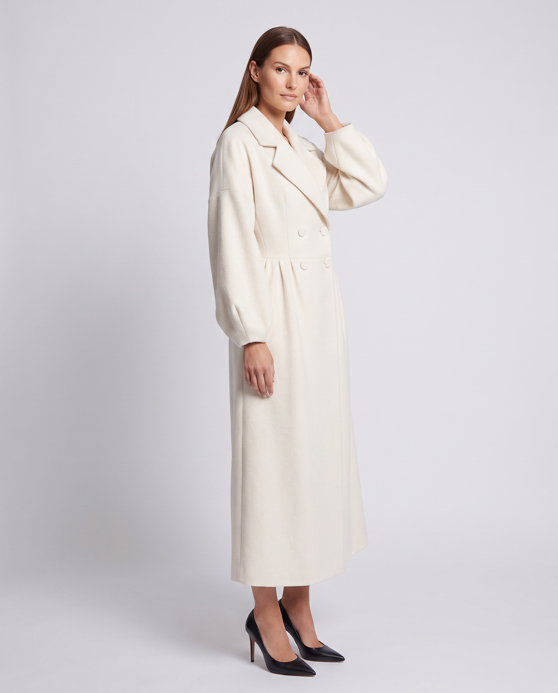 Woman wearing white double breasted maxi coat with long sleeves and black pointed toe heels, standing against light gray background