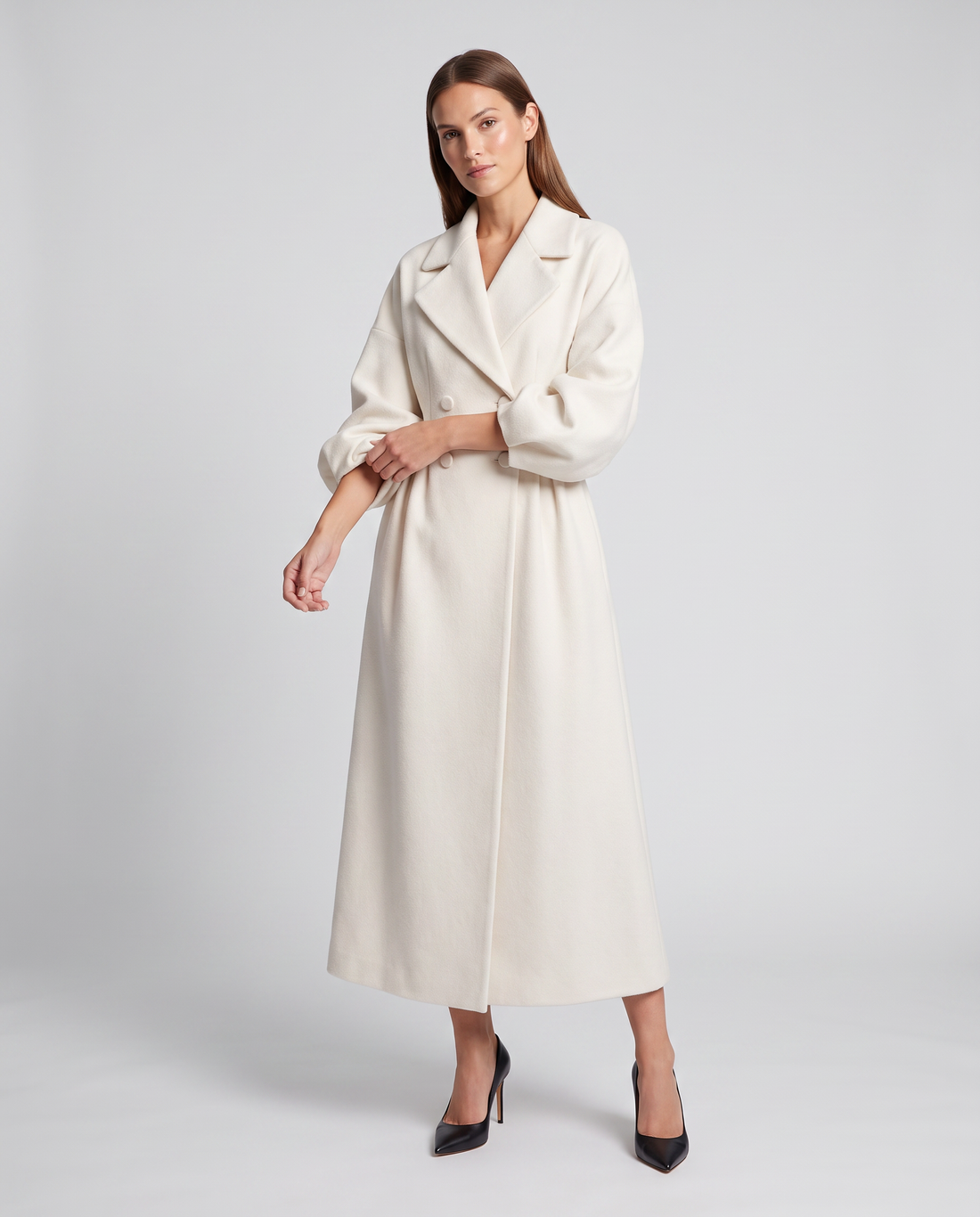 Woman wearing white double breasted maxi coat with long sleeves and black pointed heel shoes, standing against light gray background