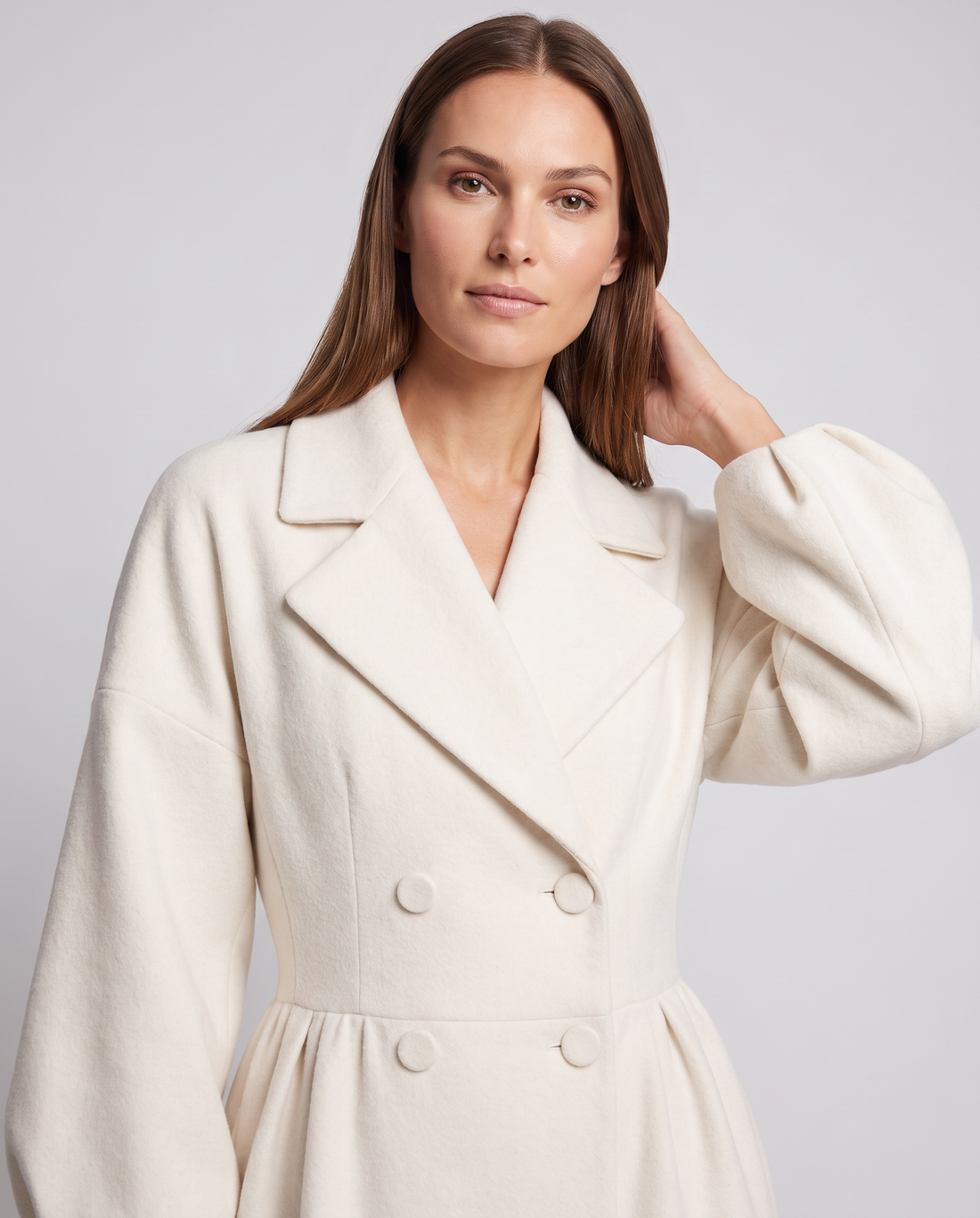 Woman wearing white double-breasted maxi coat with puffed sleeves and notch collar, hand touching hair against neutral background