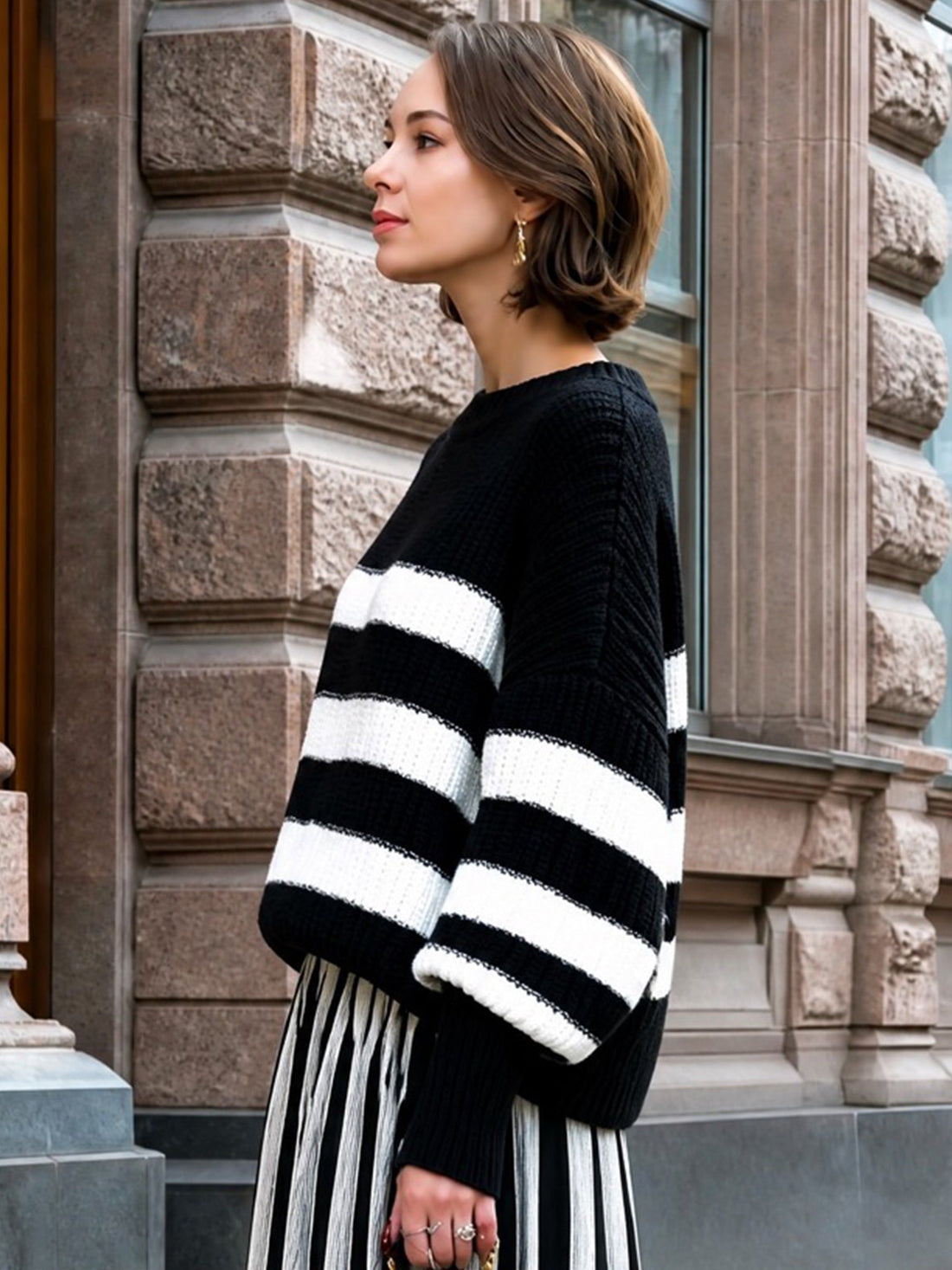 Woman wearing black and white striped relaxed knit sweater with pleated skirt, standing against stone building exterior