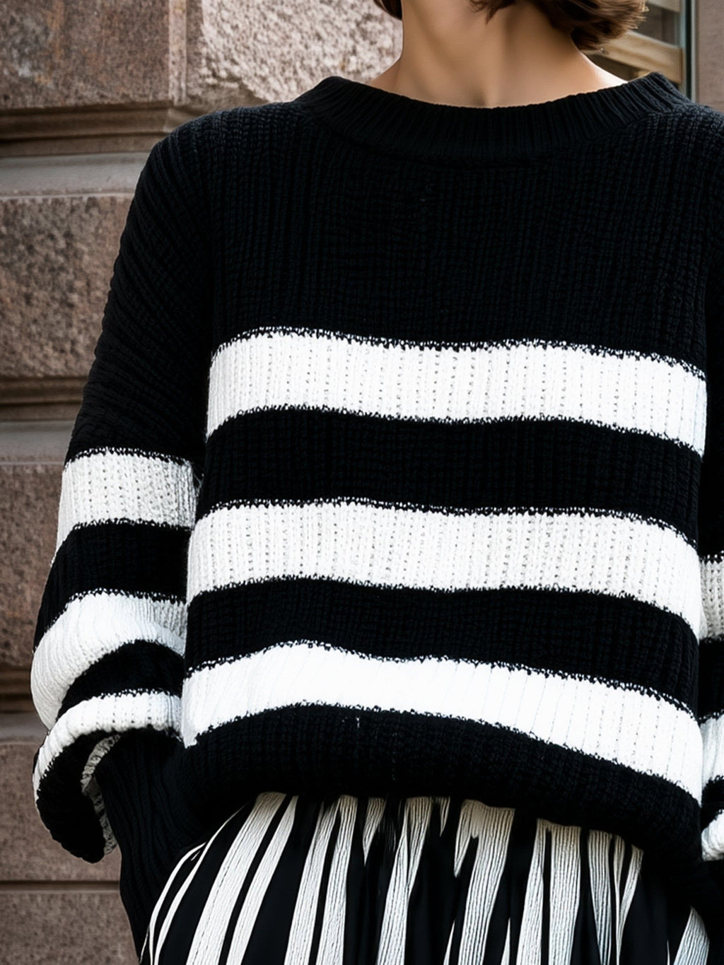 Woman wearing black and white striped relaxed knit sweater with ribbed texture against stone wall backdrop