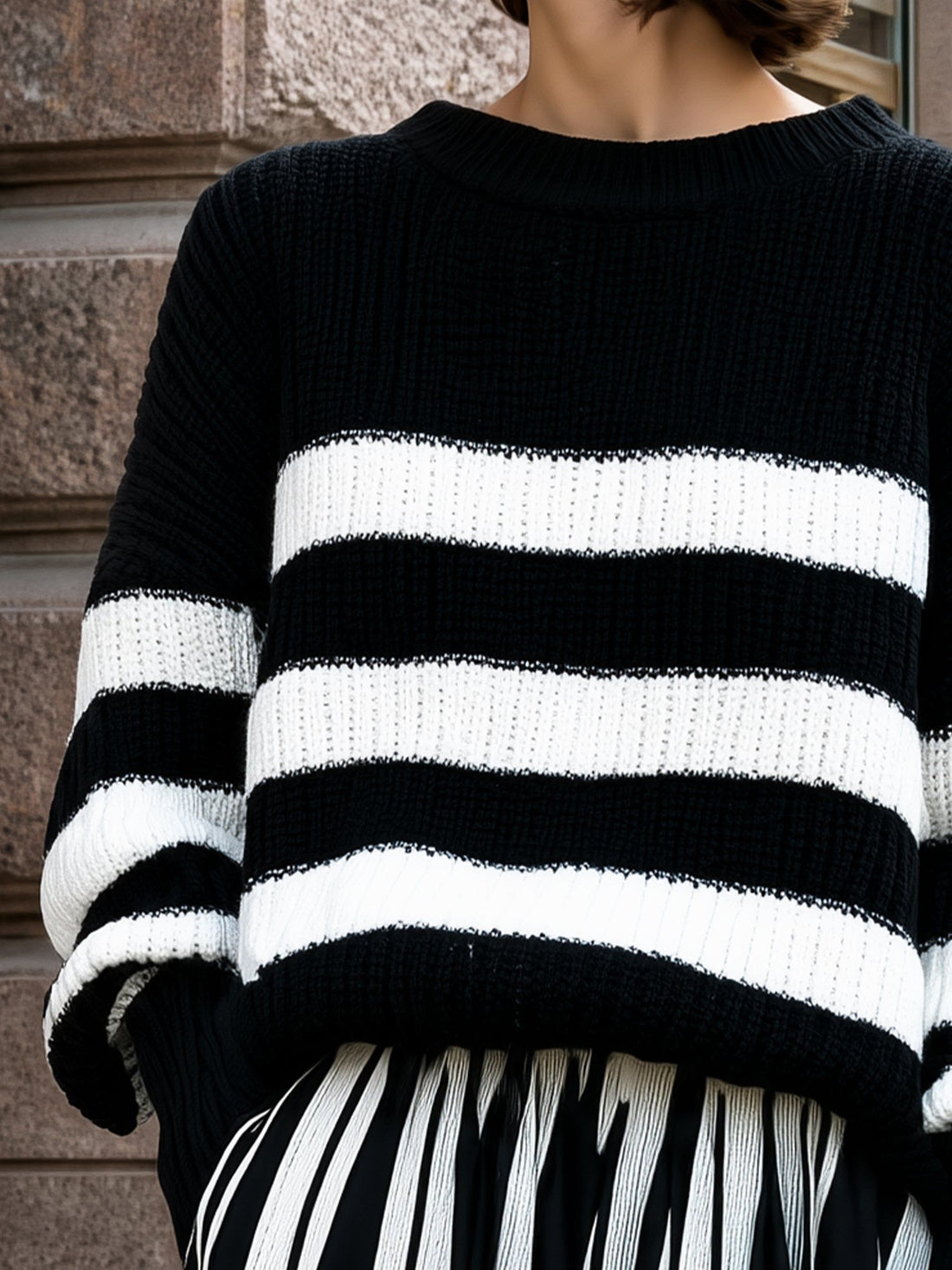 Woman wearing black and white striped relaxed knit sweater with ribbed texture against stone wall backdrop