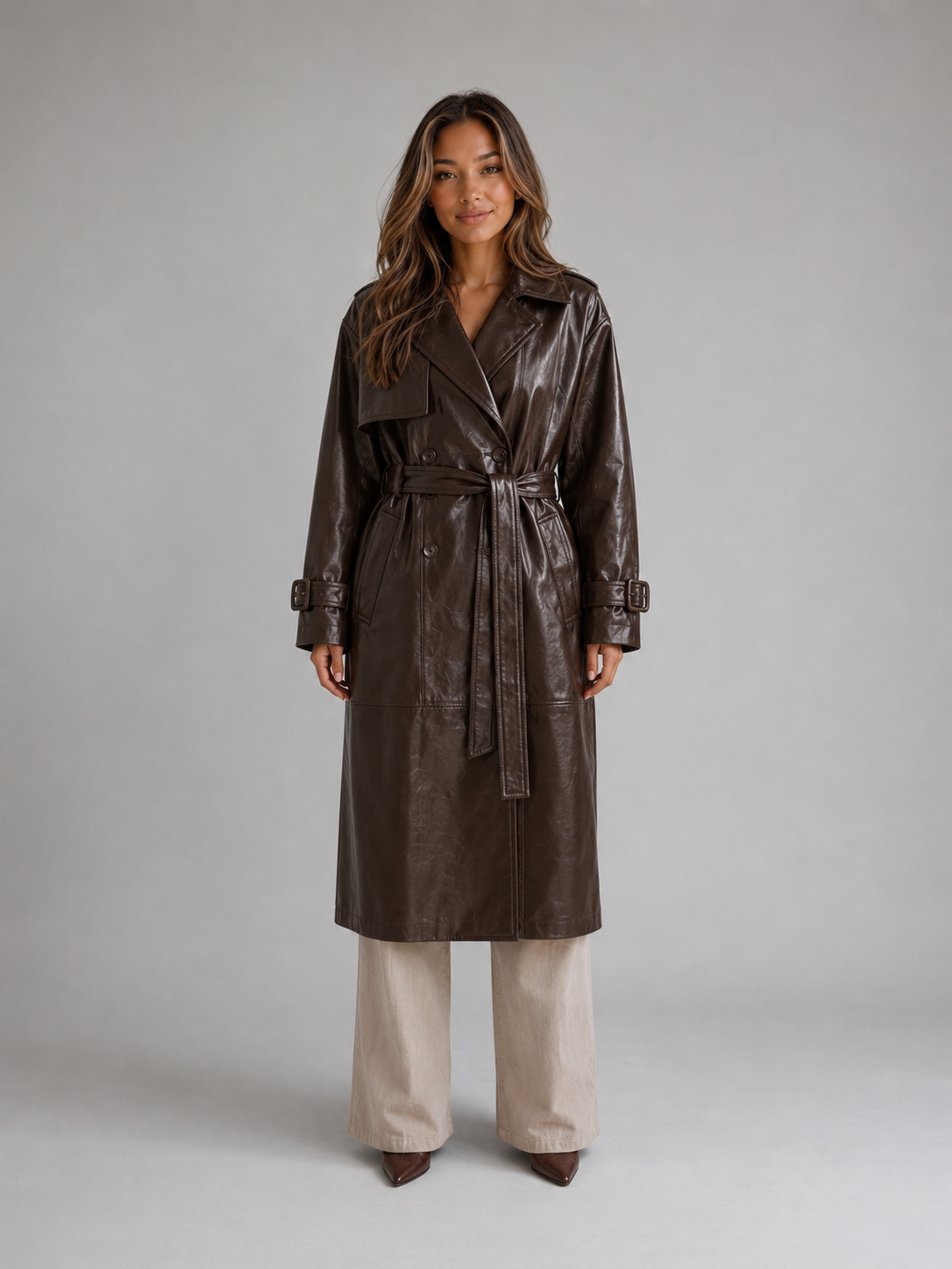 Woman wearing a long double breasted belted brown leather trench coat with cream pants and brown pointed toe heels