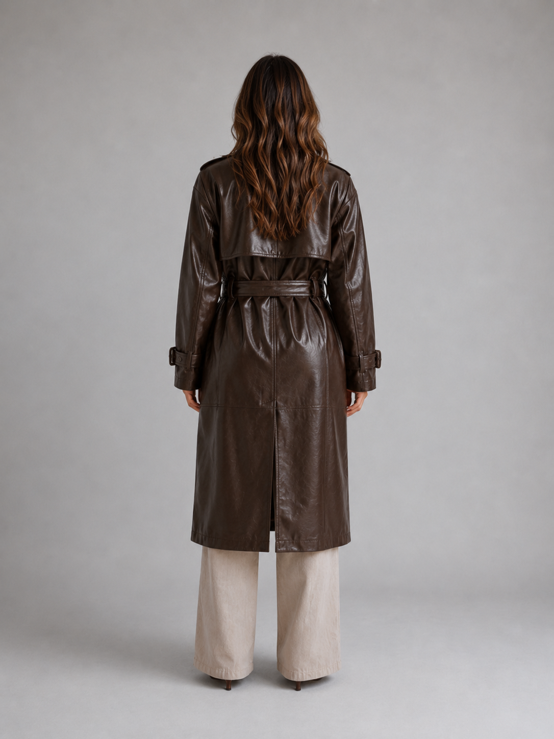 Woman wearing brown belted leather trench coat with long sleeves over cream wide-leg pants, viewed from behind