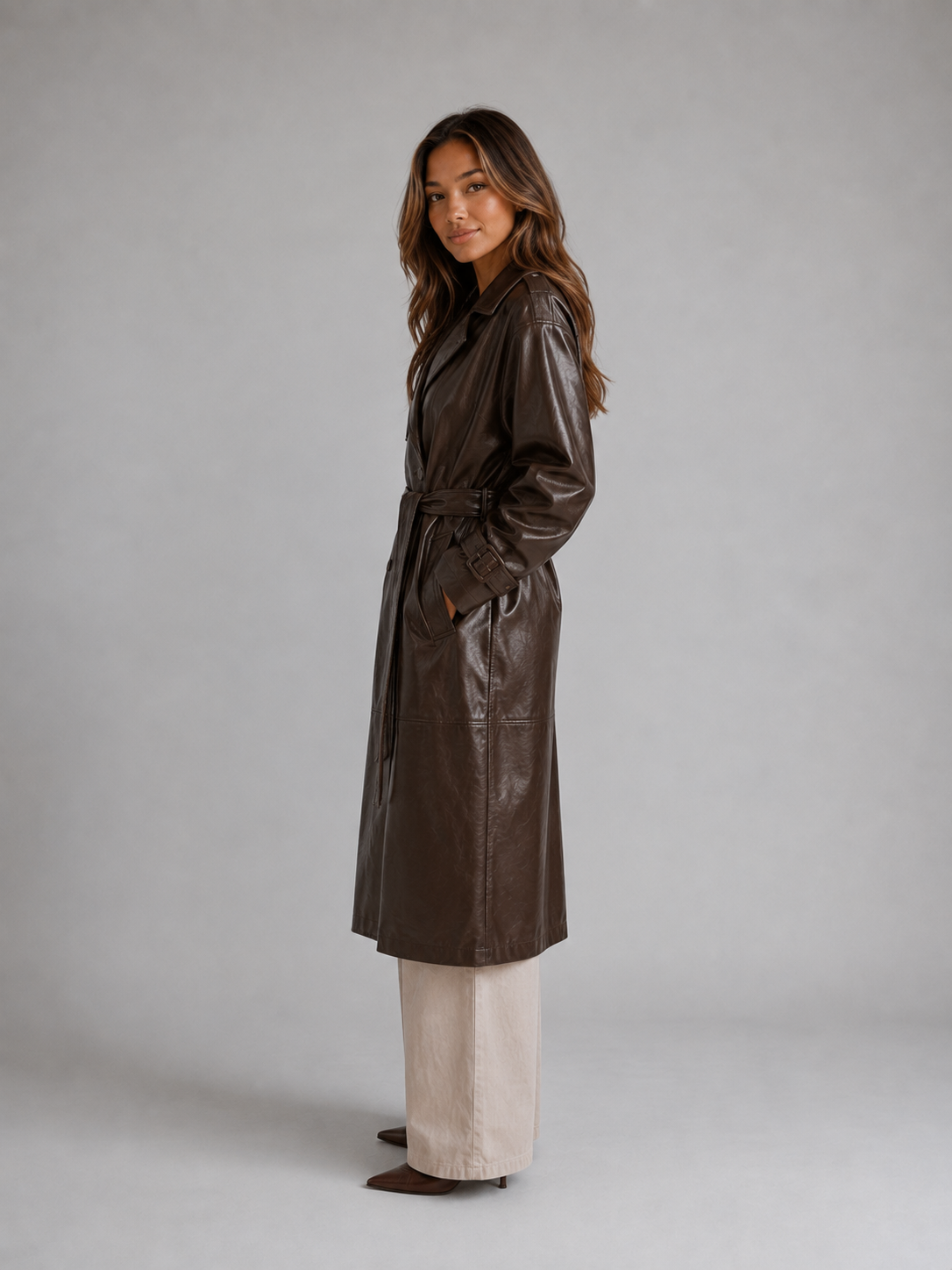 Woman wearing brown belted leather trench coat over cream pants, standing against gray background