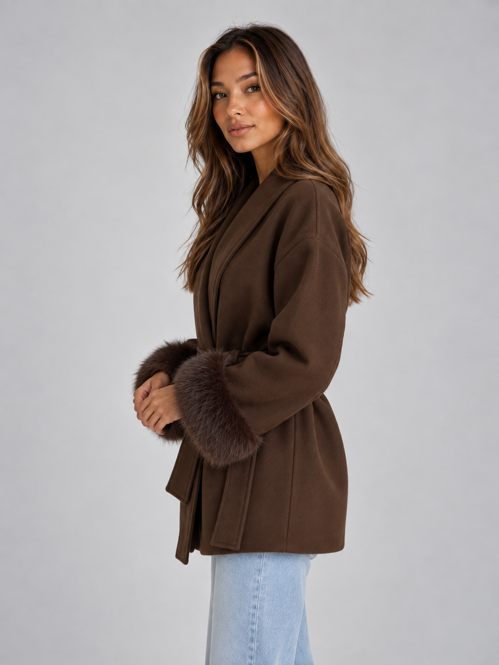 Woman wearing brown belted wrap coat with faux fur cuffs and light blue jeans, standing against white background