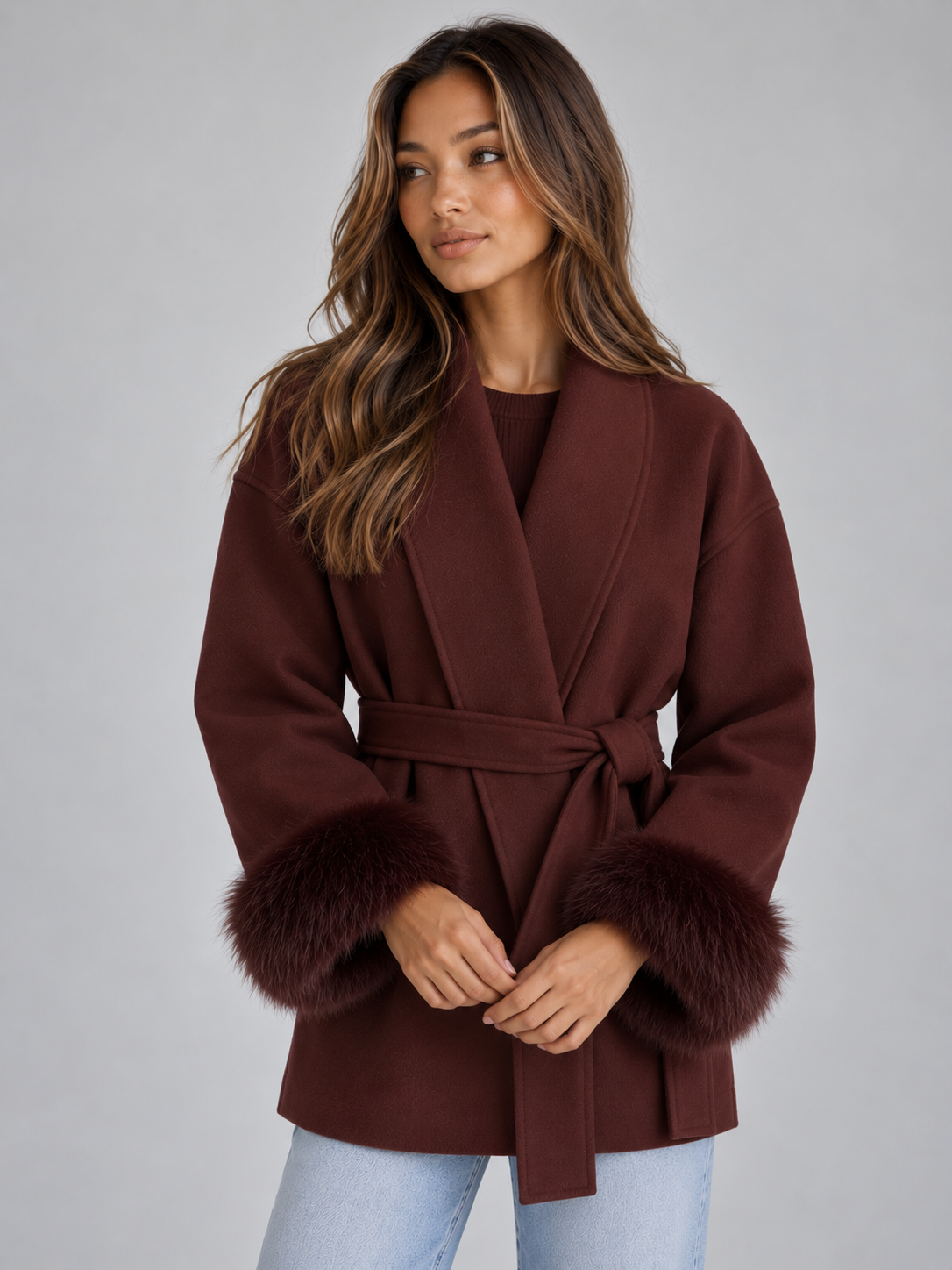 Woman wearing burgundy belted wrap coat with faux fur cuffs and light blue jeans against gray background