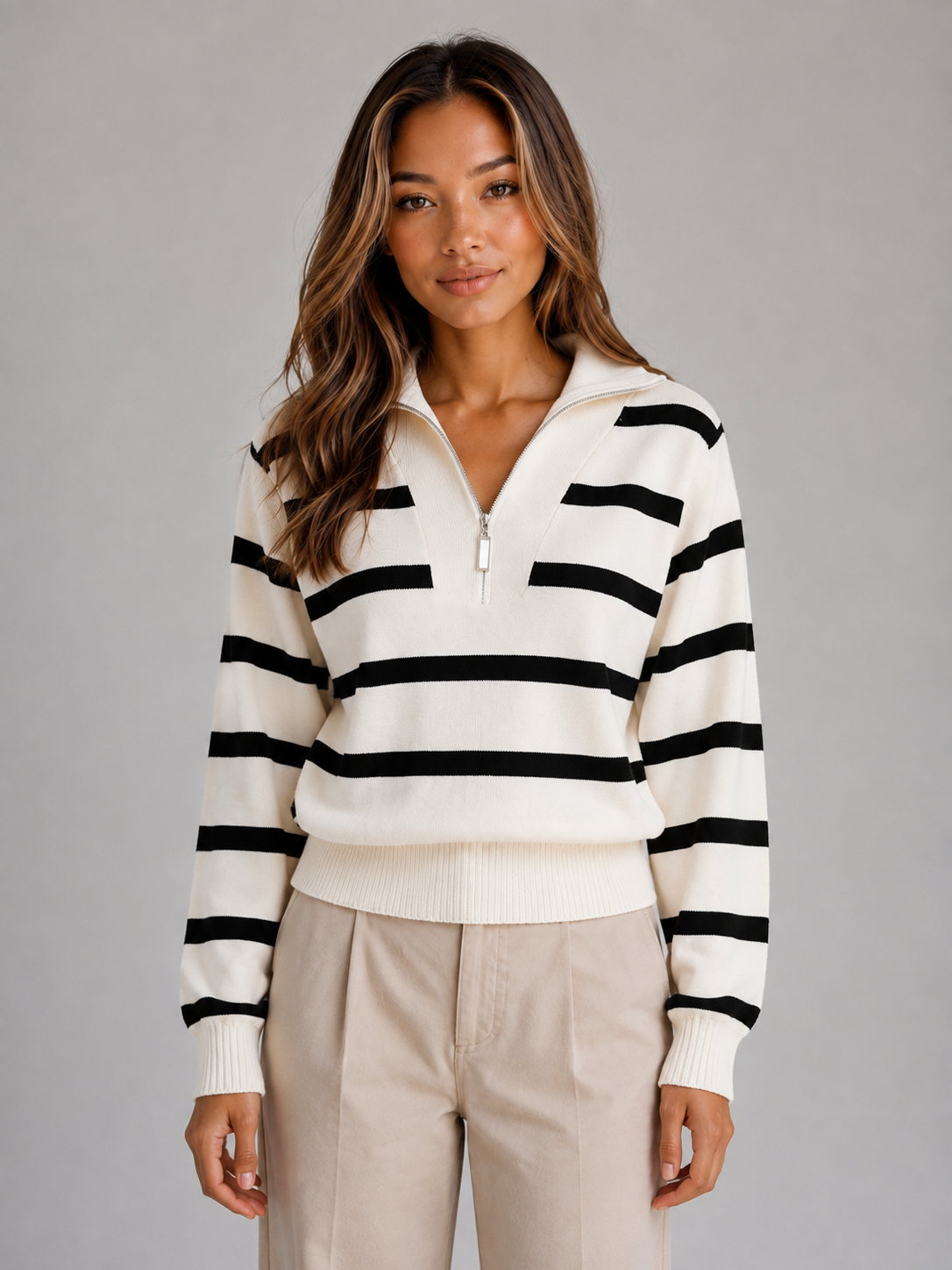 Woman wearing cream and black striped half-zip collared knit sweater with beige pants against gray background