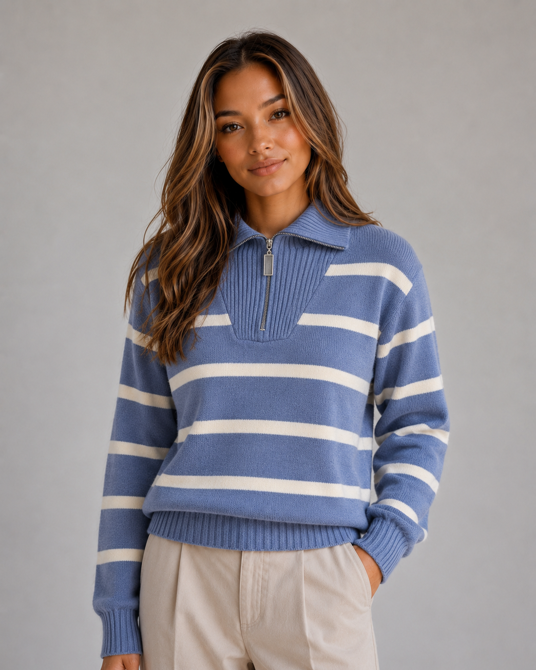 Woman wearing blue and cream striped half-zip knitted jumper with relaxed collar and beige pants against gray background
