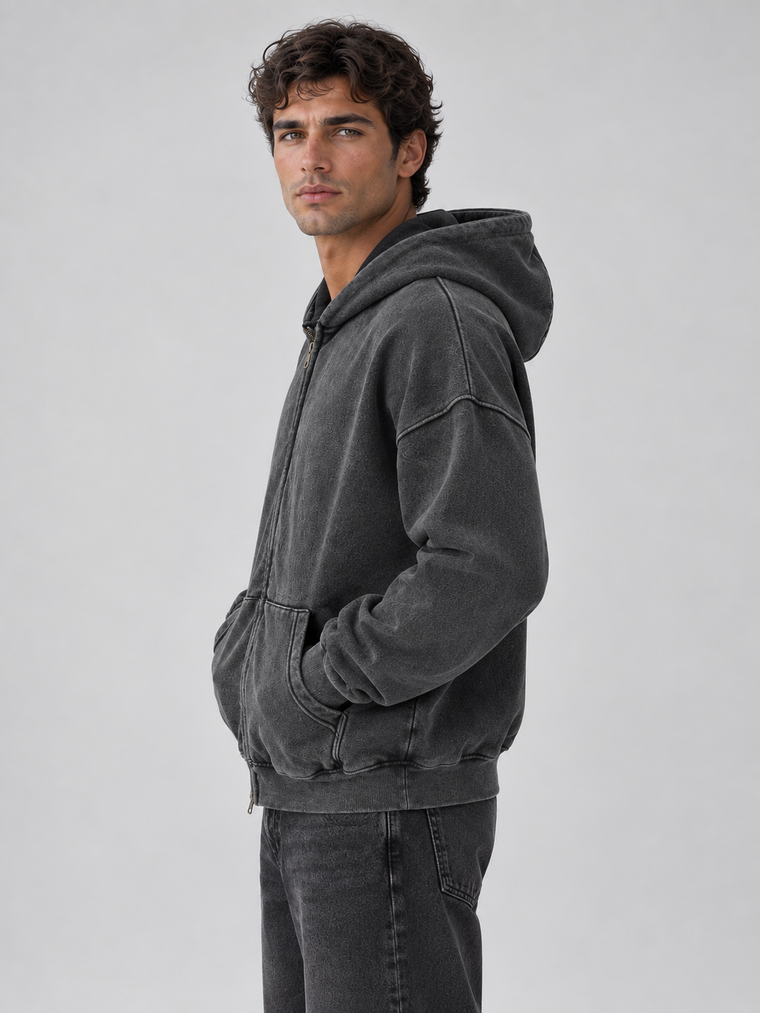 Man wearing dark gray washed oversized hooded zip-up jacket with denim jeans, shown from side profile against light background