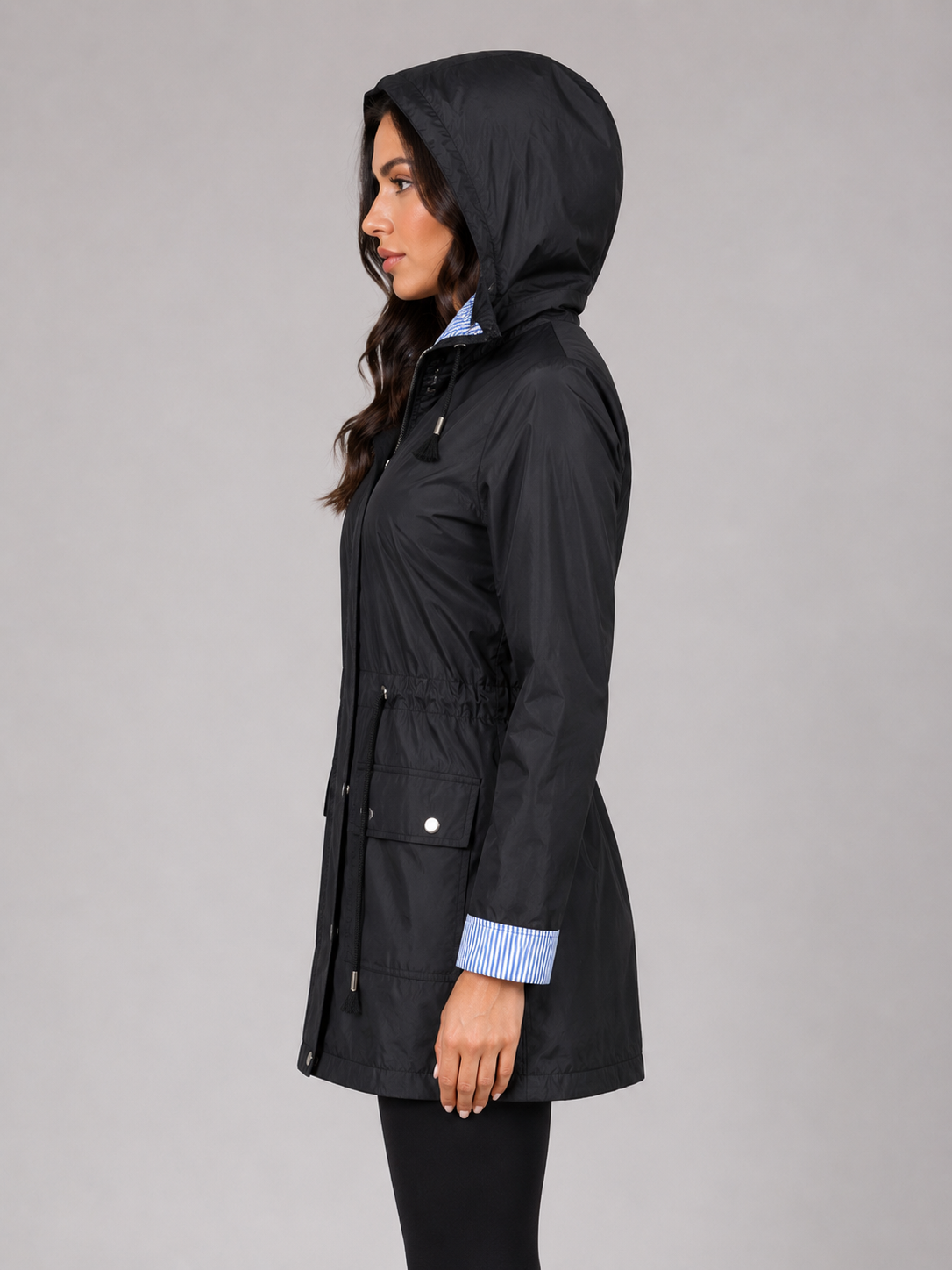 Woman wearing black lightweight water resistant hooded rain jacket with blue striped shirt cuffs visible, shown in profile view against gray background