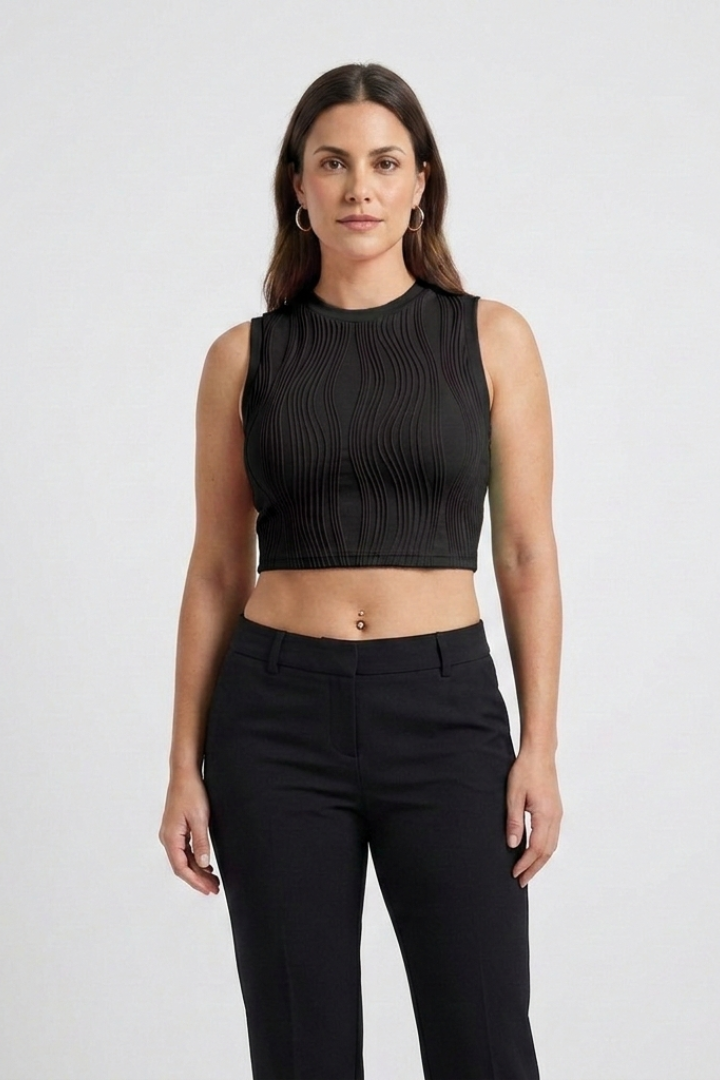 Woman wearing black ribbed sleeveless crop top with high neckline and black trousers, standing against white background