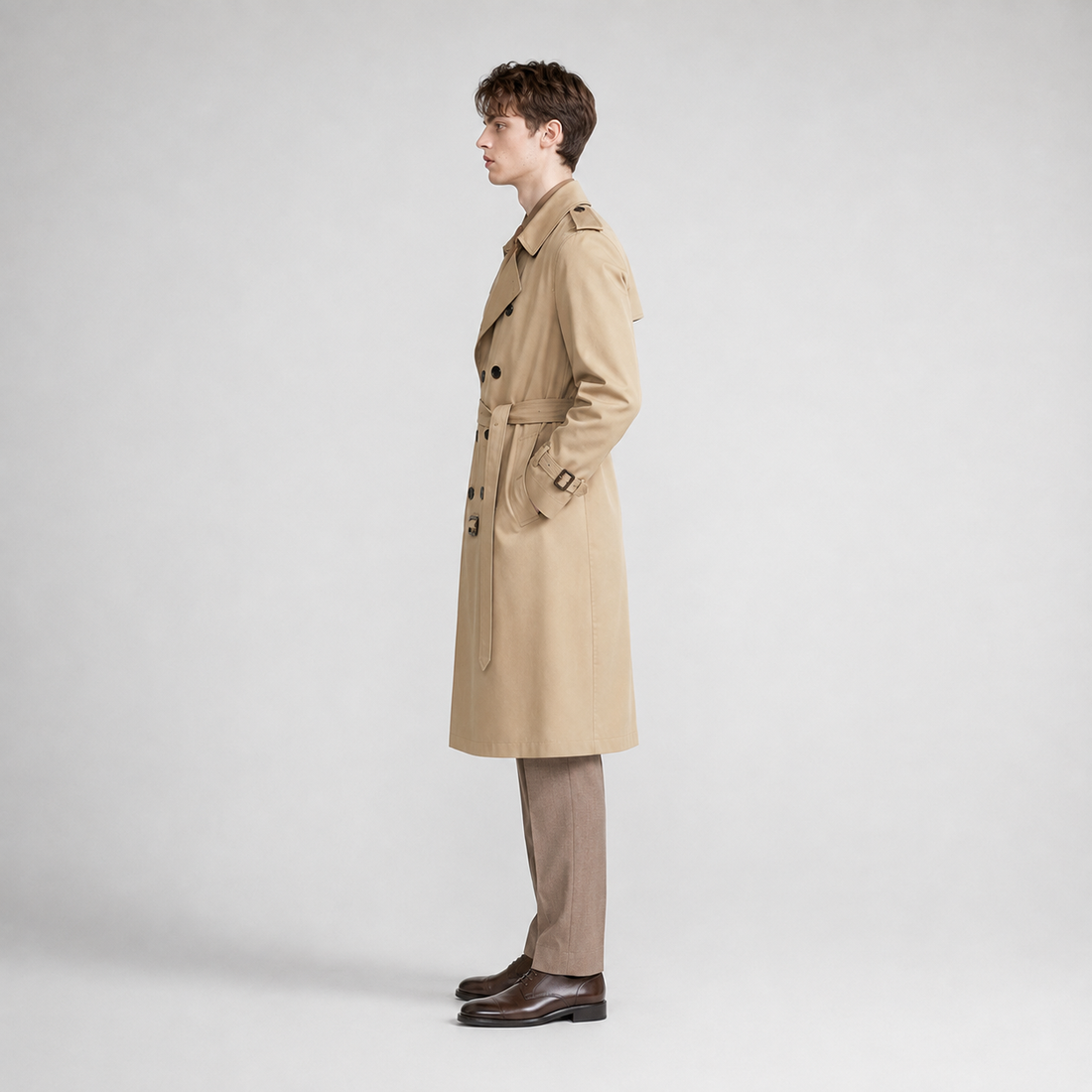 Man wearing a beige belted double breasted trench coat with long sleeves over tan pants and brown leather shoes, standing in profile against a white background