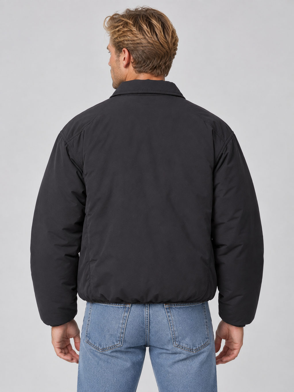 Man wearing black lightweight padded collared bomber jacket with full zip over blue jeans, back view