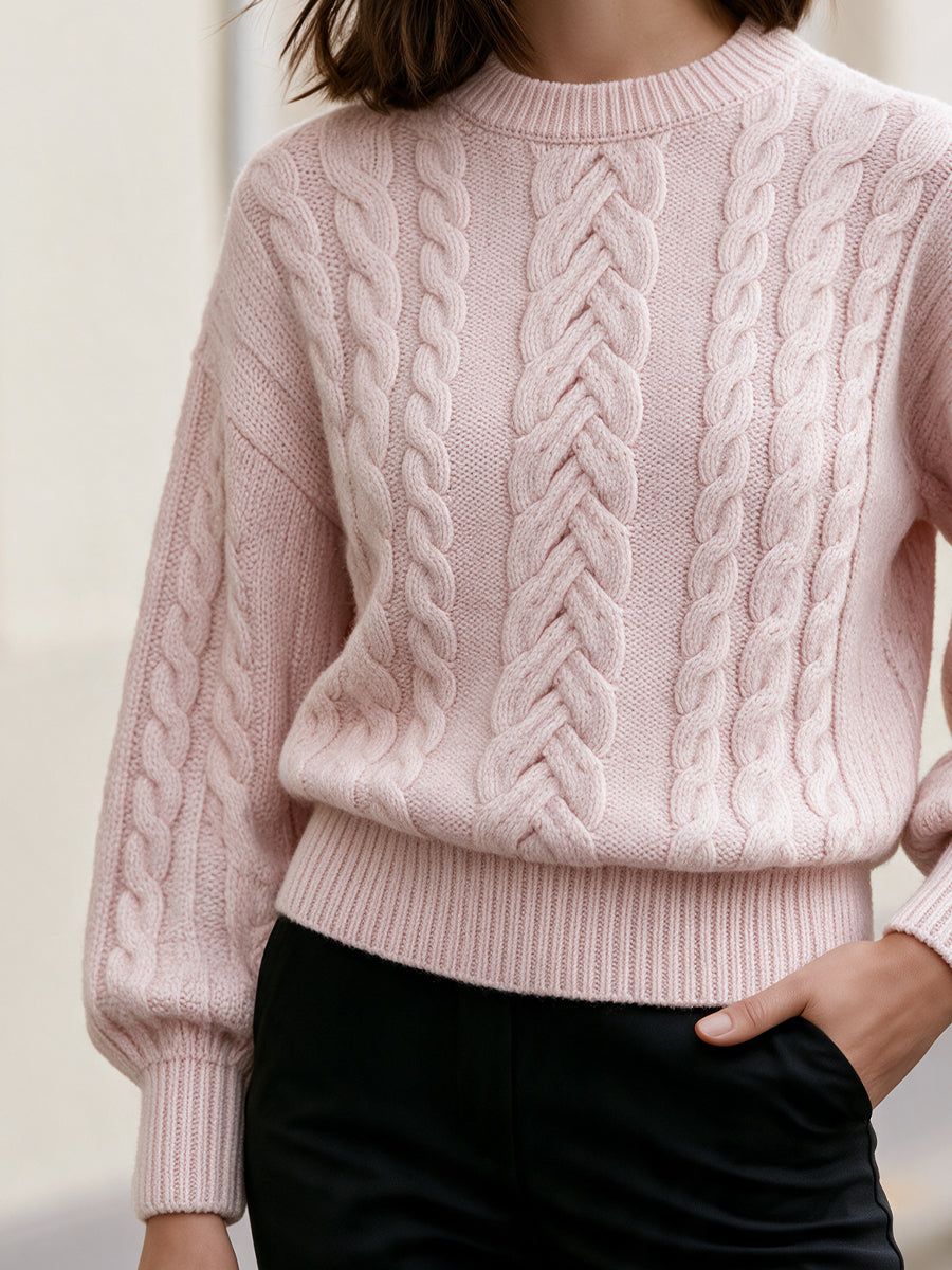 Woman wearing a light pink cable-knit cropped sweater with drop shoulders and ribbed cuffs, paired with black pants