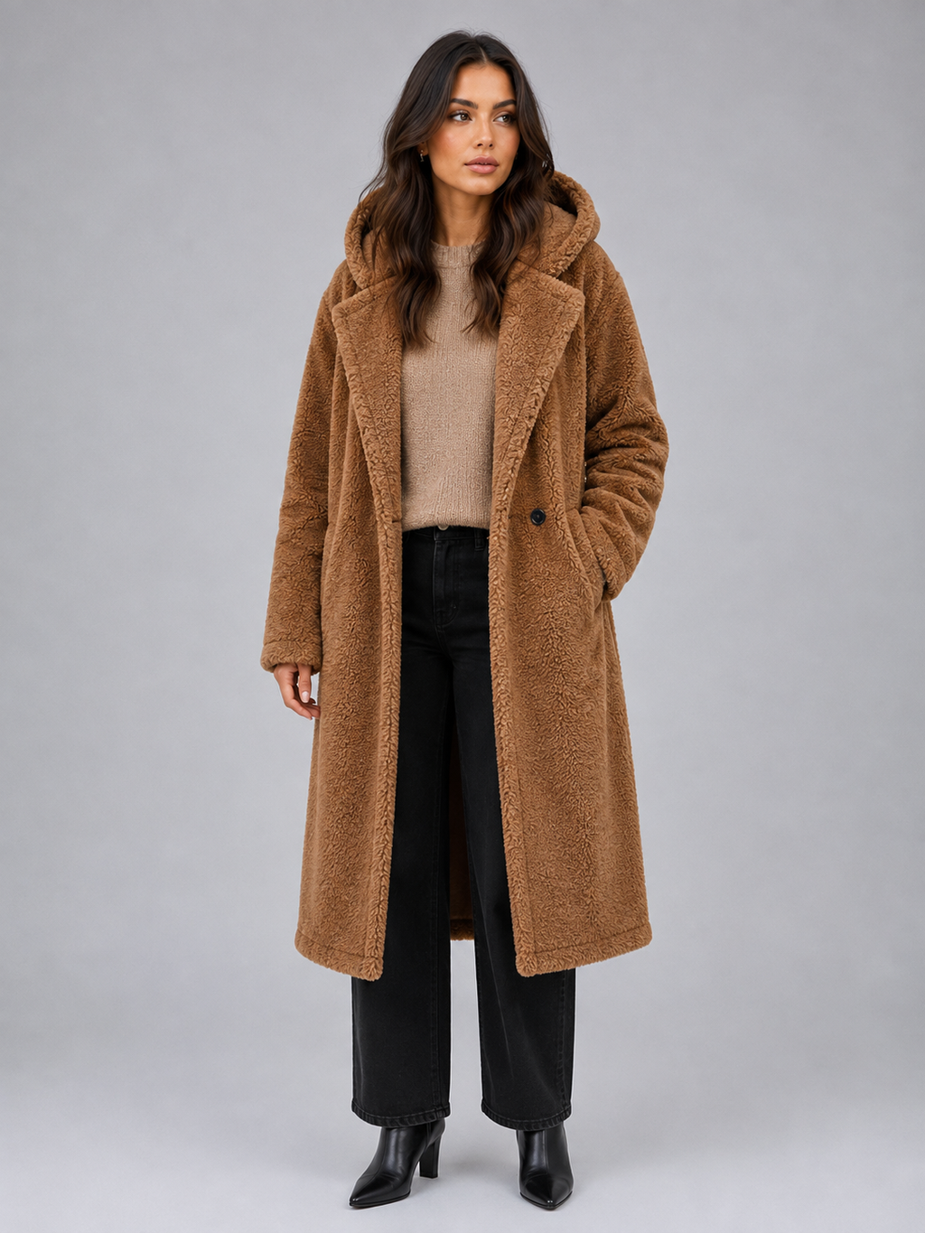 Woman wearing camel teddy coat with hood over beige sweater, black jeans, and black boots against gray background