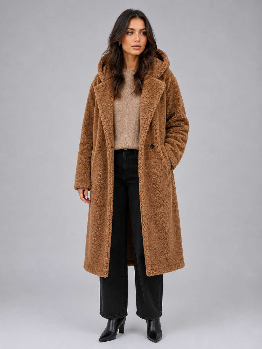 Woman wearing camel teddy coat with hood over beige sweater, black jeans, and black boots against gray background