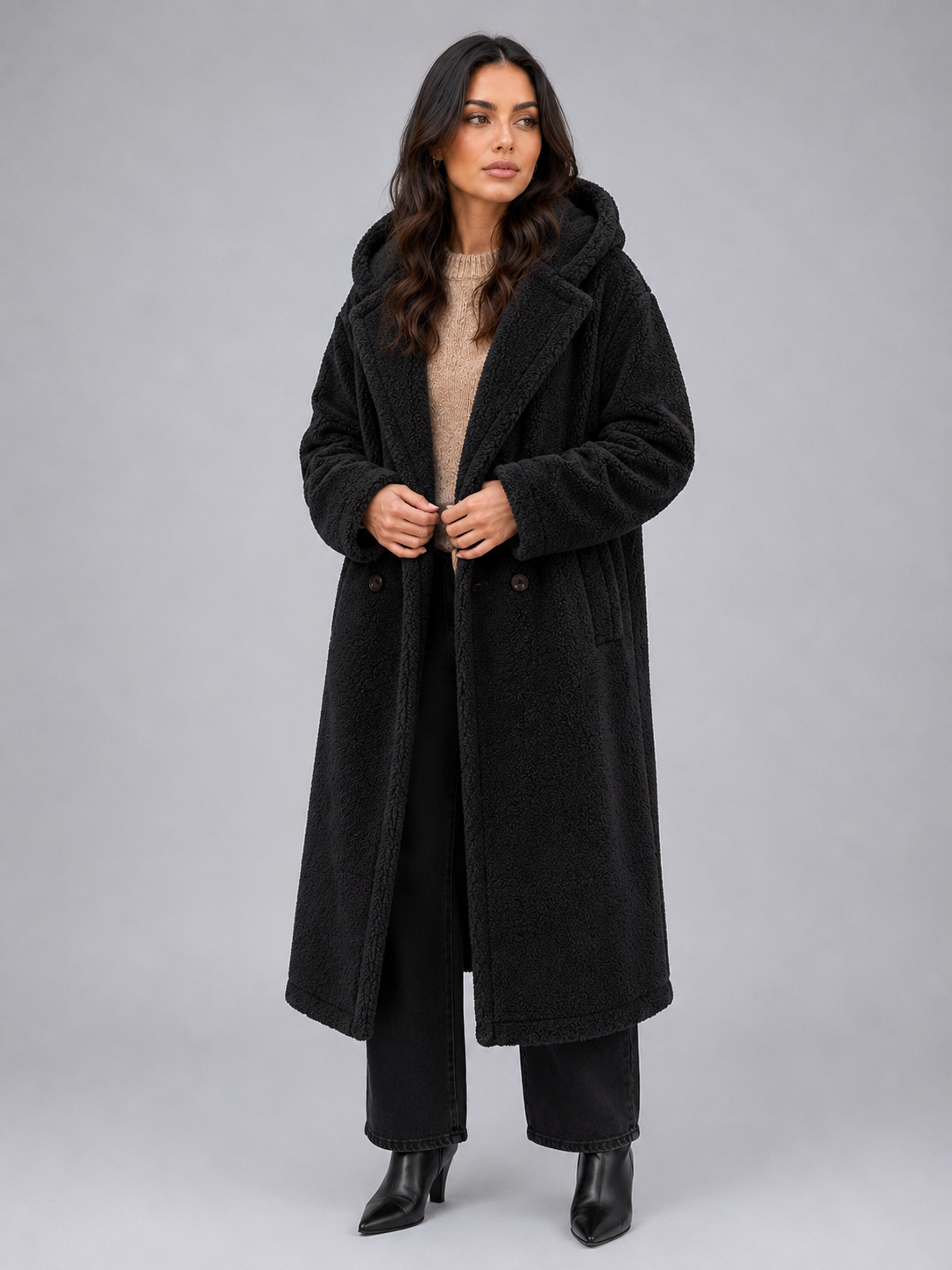 Woman wearing black oversized hooded borg teddy coat with beige sweater, black jeans and black boots against gray background