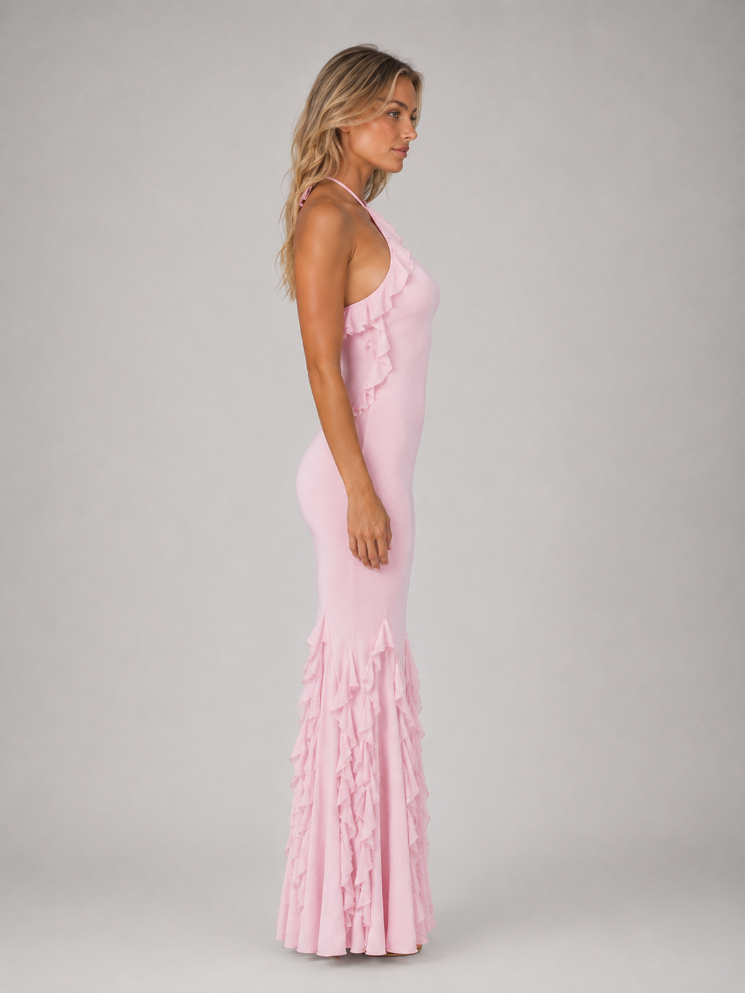Woman wearing pink halterneck bodycon maxi gown with ruffled fishtail hem, side profile view against grey background