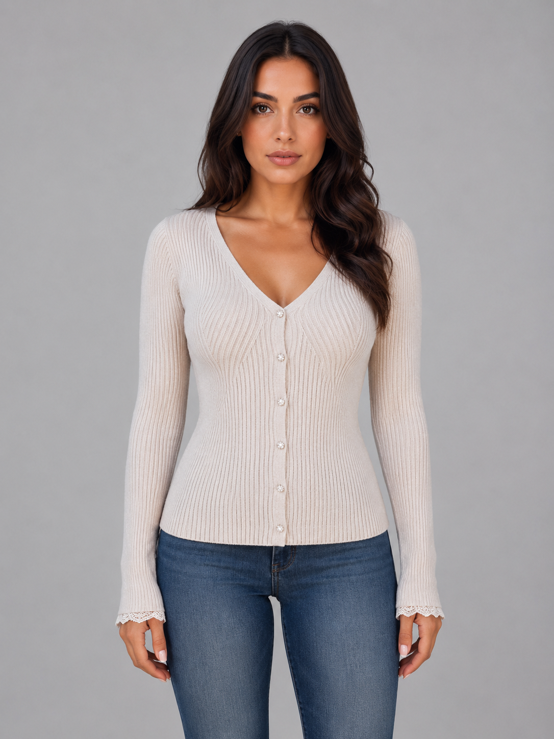 Woman wearing cream ribbed knit cardigan with buttons and lace cuffs over blue jeans, gray background