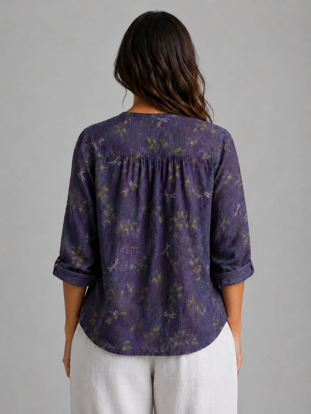 Woman wearing purple floral relaxed v-neck blouse with rolled sleeves and white pants, back view