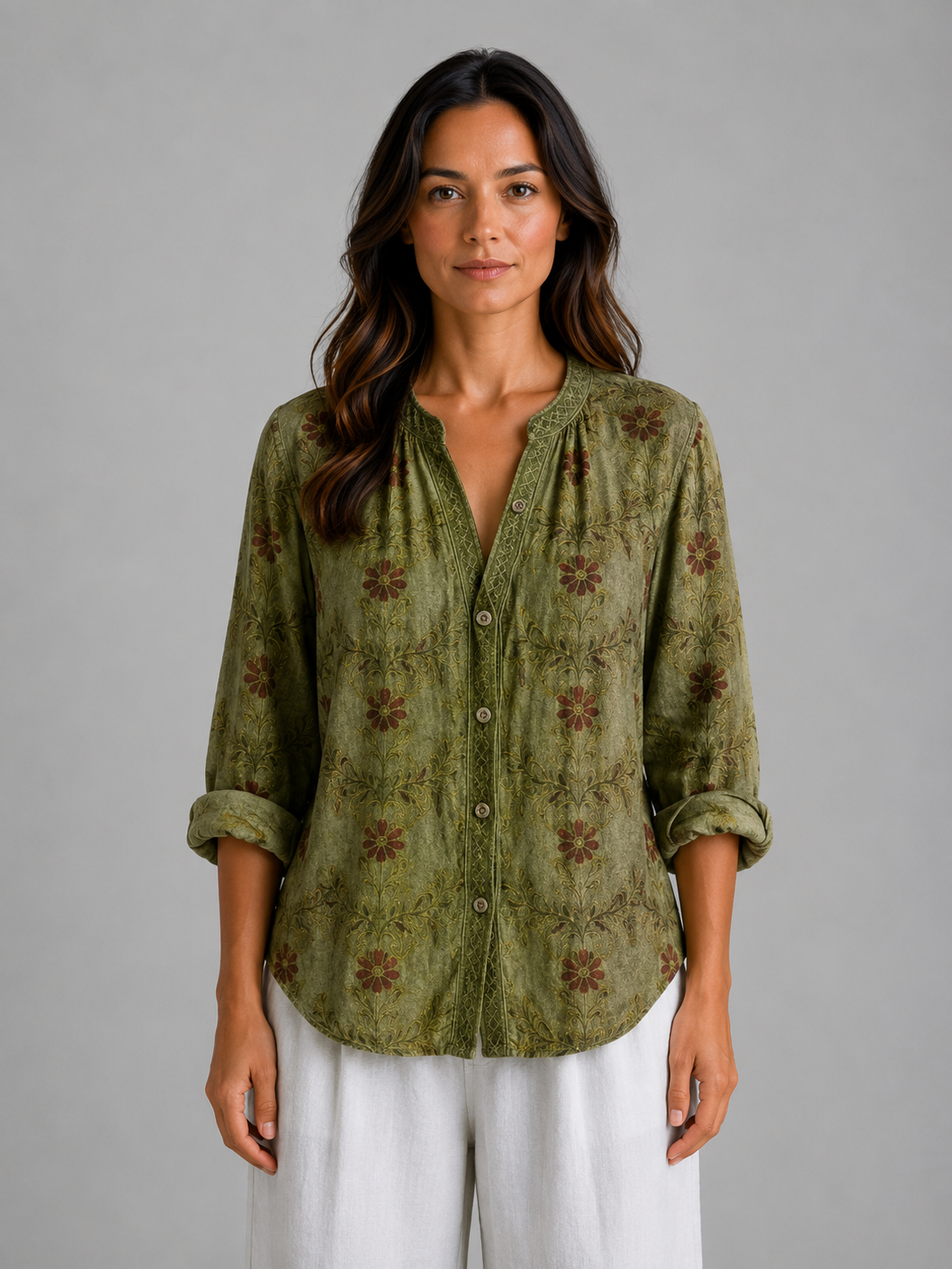 Woman wearing olive green floral print v-neck blouse with rolled sleeves and white pants, standing against gray background