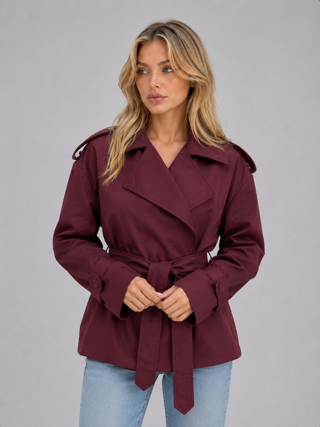 Woman wearing burgundy belted double breasted short trench jacket with light blue jeans, neutral background