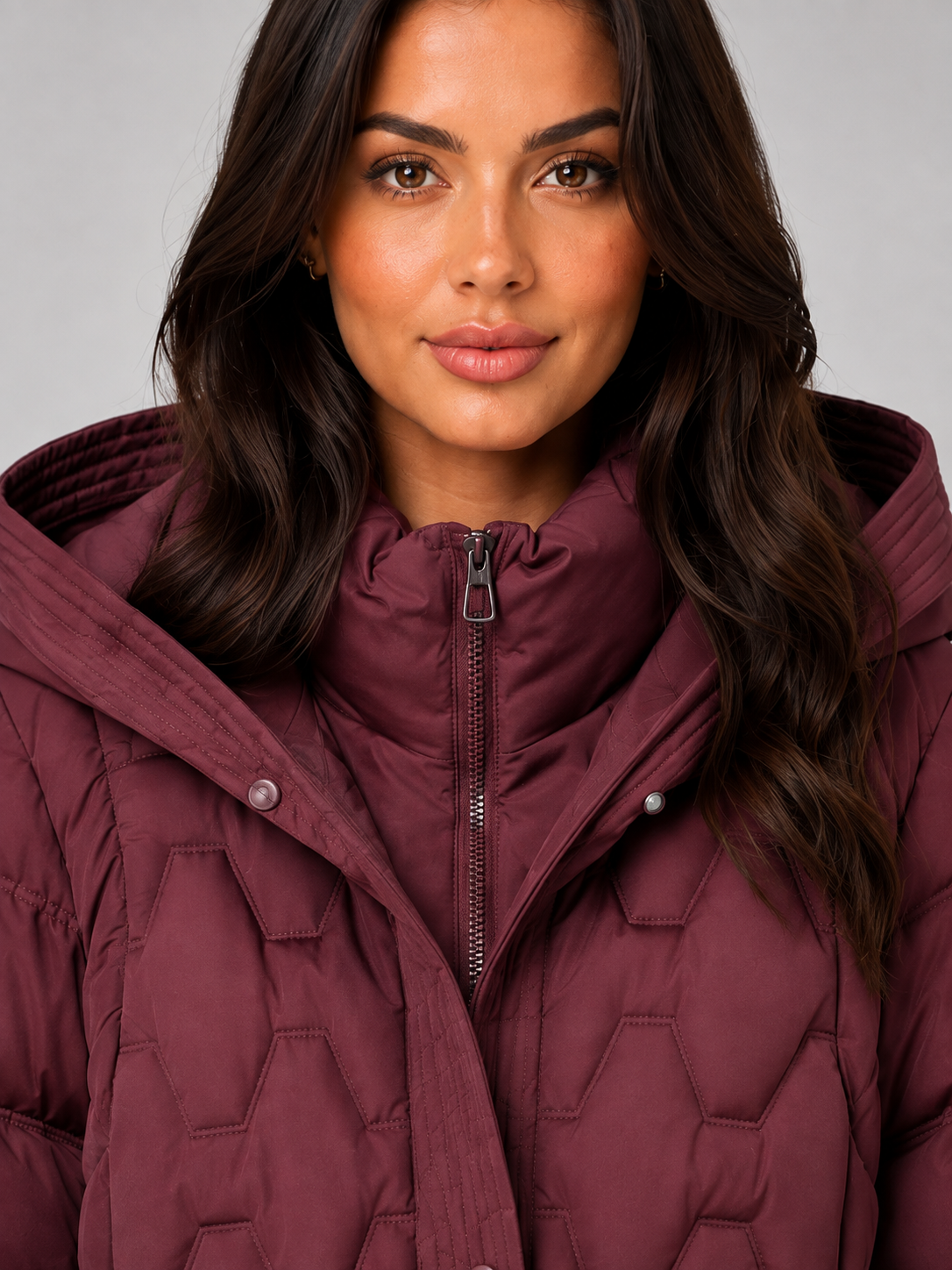 Woman wearing maroon quilted hooded long puffer coat with zip front and chest pockets against gray background