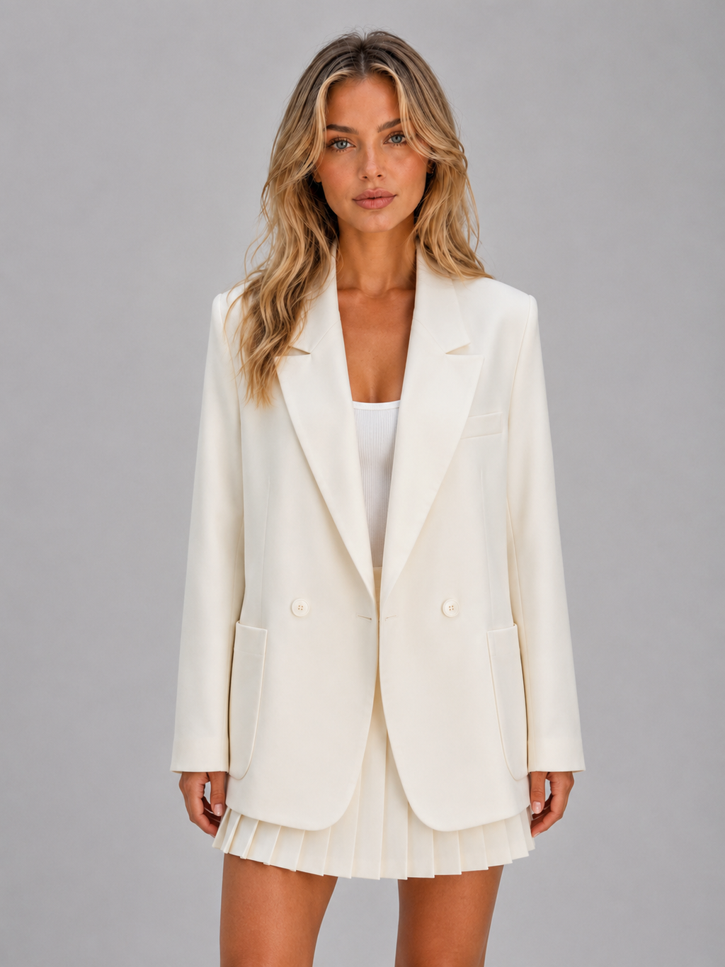 Woman wearing cream double-breasted oversized blazer with white ribbed top and pleated mini skirt on gray background