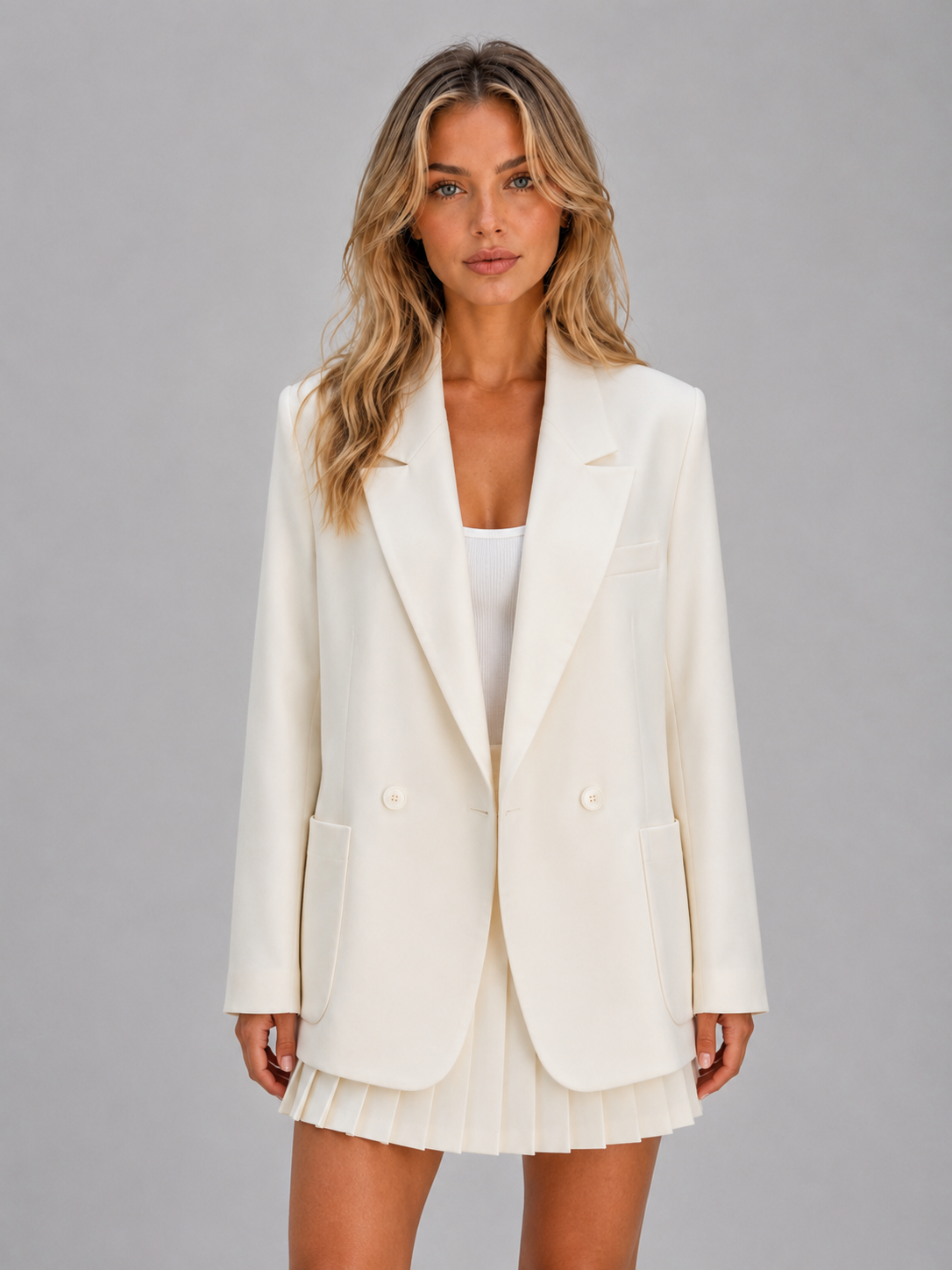 Woman wearing cream double-breasted oversized blazer with white ribbed top and pleated mini skirt on gray background