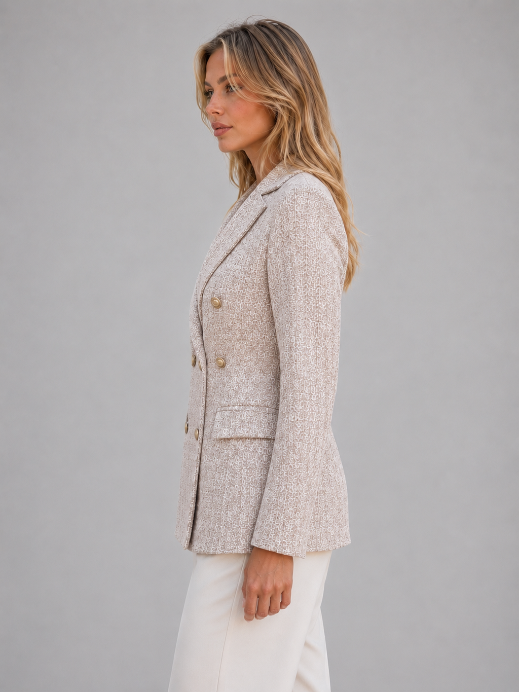 Woman wearing beige tweed double-breasted blazer jacket with white pants, side view against gray background
