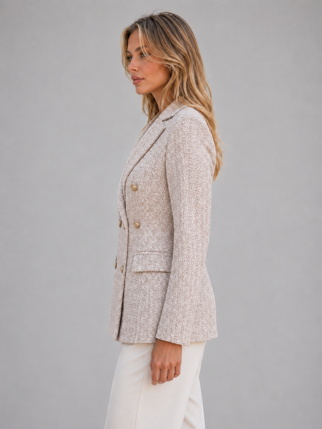 Woman wearing beige tweed double-breasted blazer jacket with white pants, side view against gray background