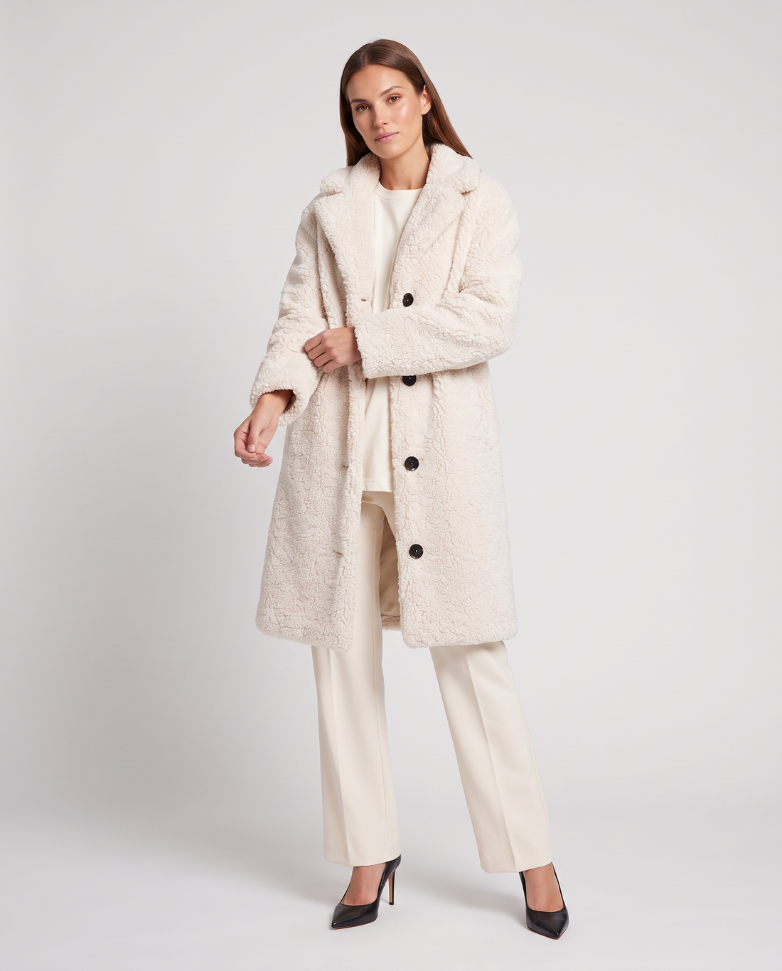 Woman wearing cream single breasted teddy coat with black buttons over white top and cream trousers, styled with black pointed heels