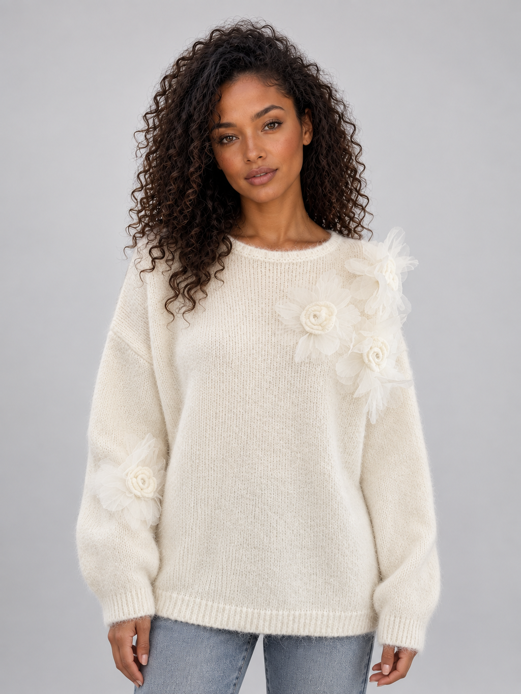 Woman wearing cream knit jumper with 3D floral details on sleeves and crew neckline, styled with blue jeans