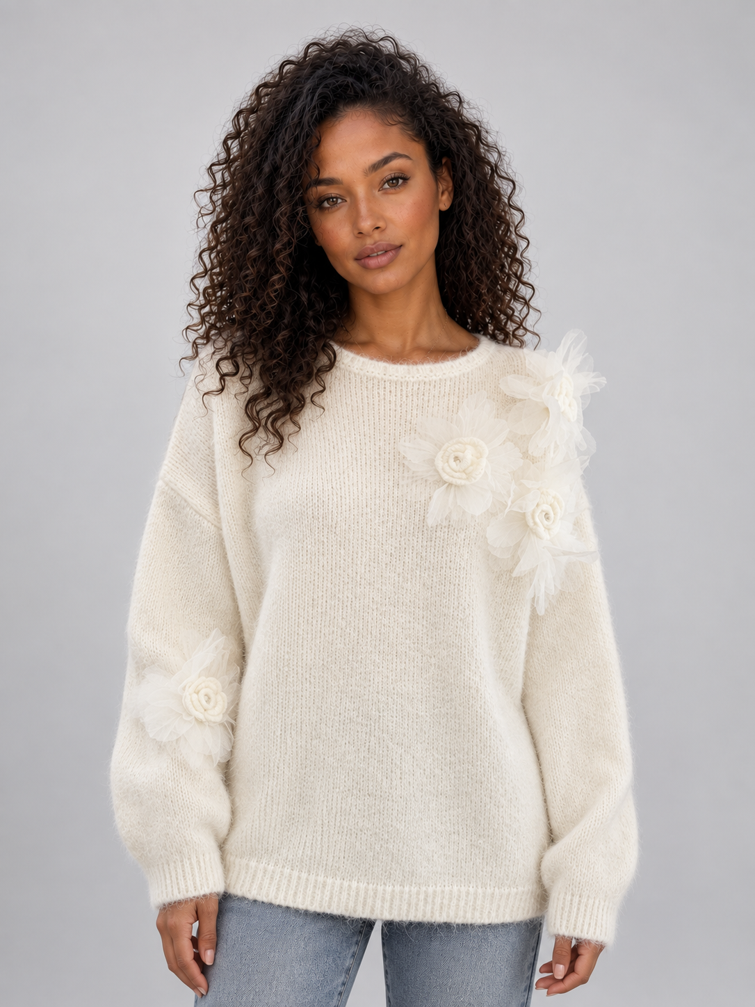 Woman wearing cream knit jumper with 3D floral details on sleeves and crew neckline, styled with blue jeans