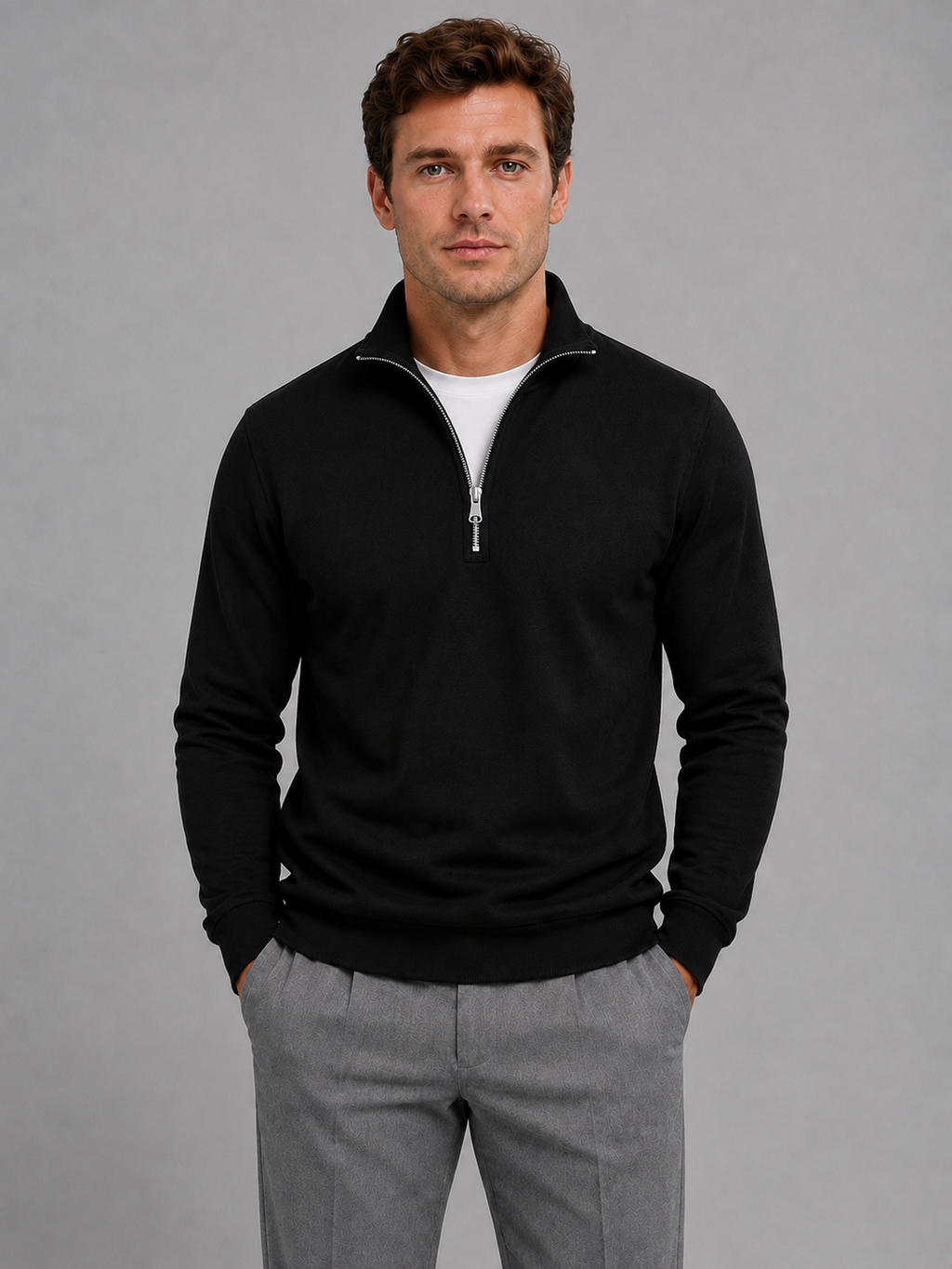 Man wearing black half-zip pullover sweatshirt with white undershirt and gray pants against gray background