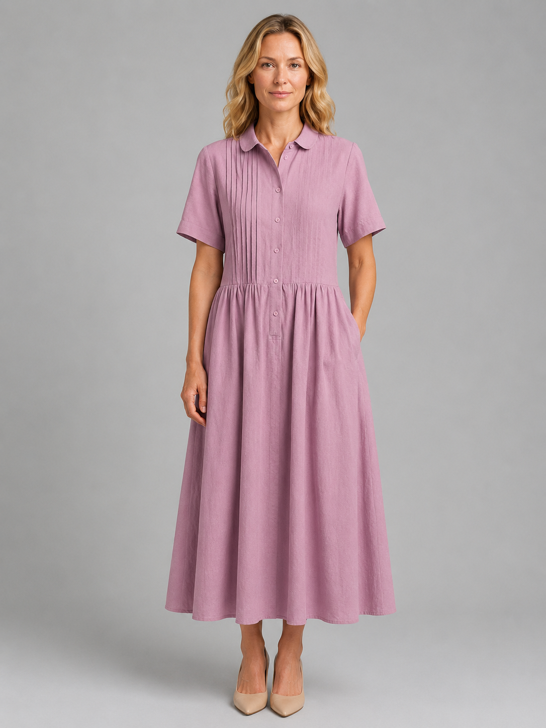 Woman wearing a pink relaxed pleated button-up maxi shirt dress with short sleeves and side pockets, paired with beige heels