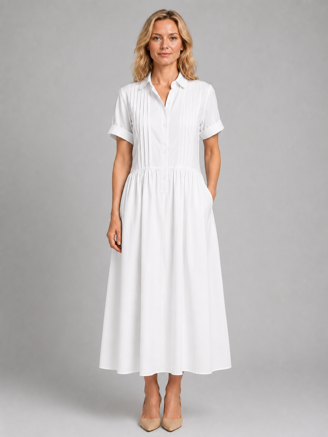 Woman wearing white short-sleeve pleated shirt maxi dress with pockets and beige heels, standing against gray background