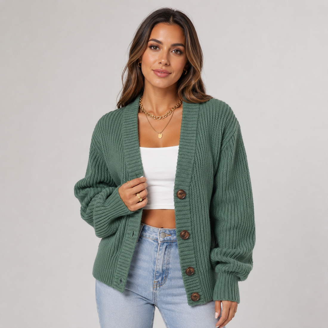 Woman wearing sage green ribbed cardigan with brown buttons over white crop top and light blue jeans, styled with gold chain necklace