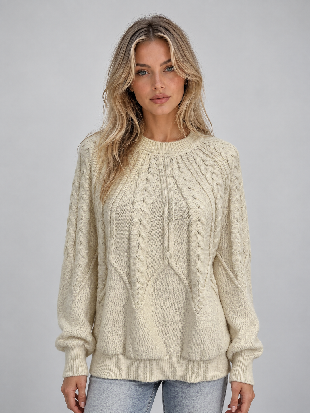 Woman wearing cream cable knit crew neck sweater with long sleeves and ribbed cuffs, paired with light blue jeans