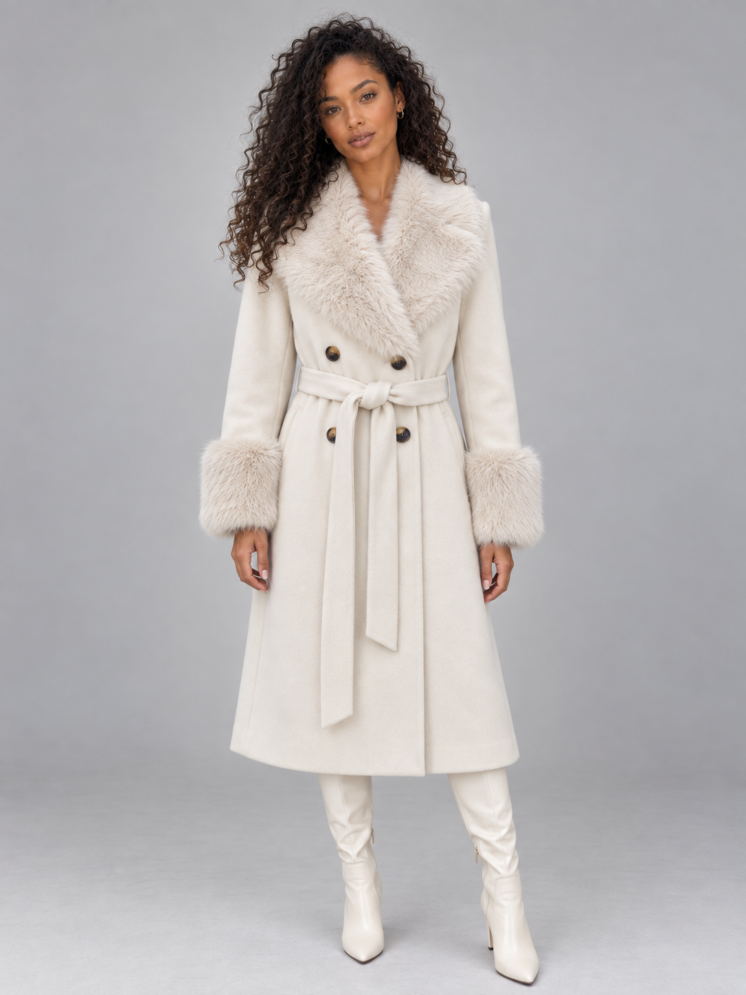 Woman wearing cream long winter coat with faux fur collar, double-breasted buttons, belt tie, and matching white knee-high boots
