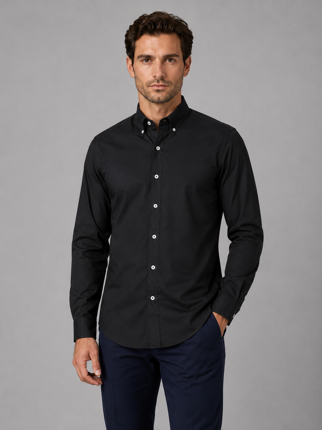 Man wearing black button down collar long sleeve shirt with white buttons and dark navy pants against gray background