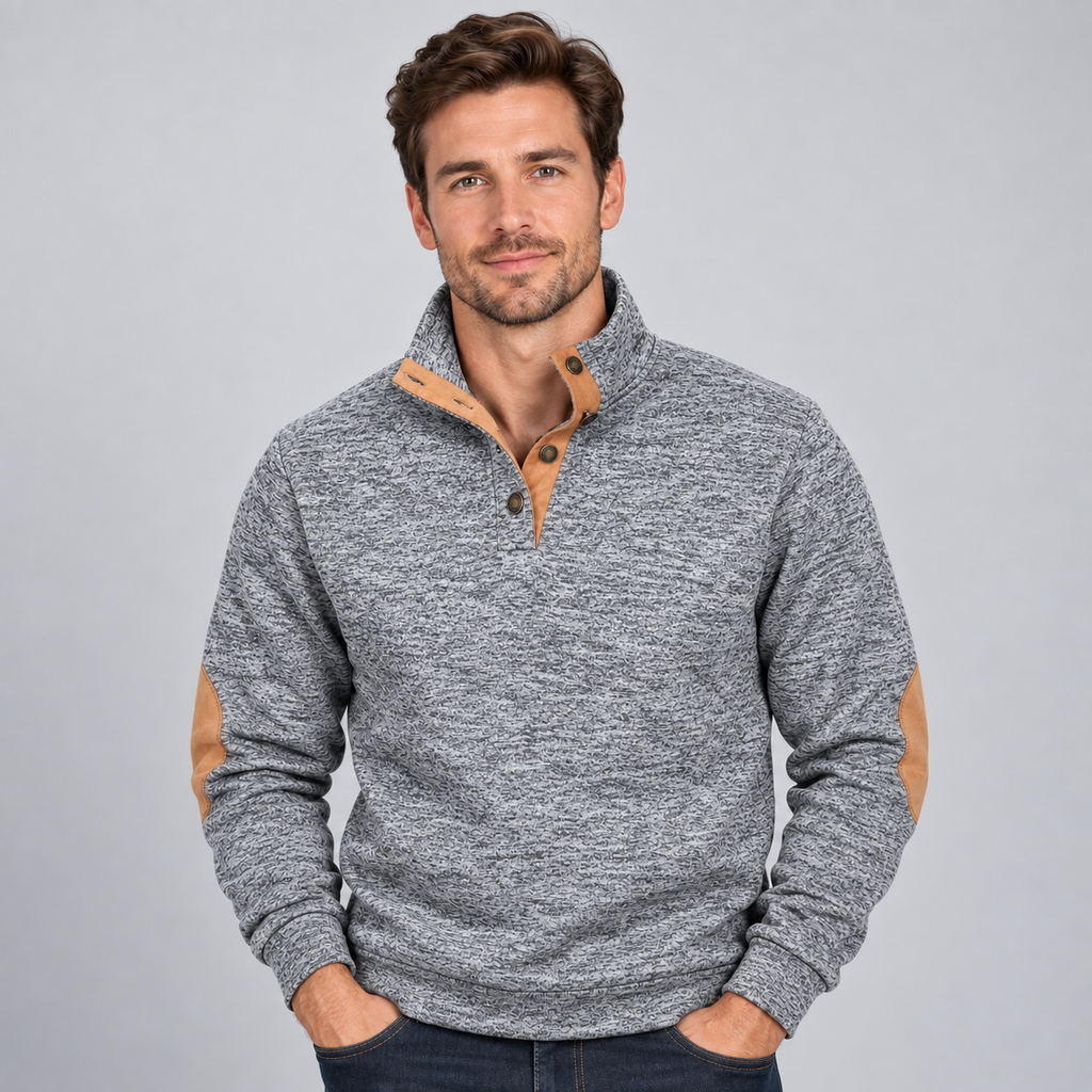 Man wearing gray quarter button stand collar jumper with tan elbow patches and dark jeans against neutral background