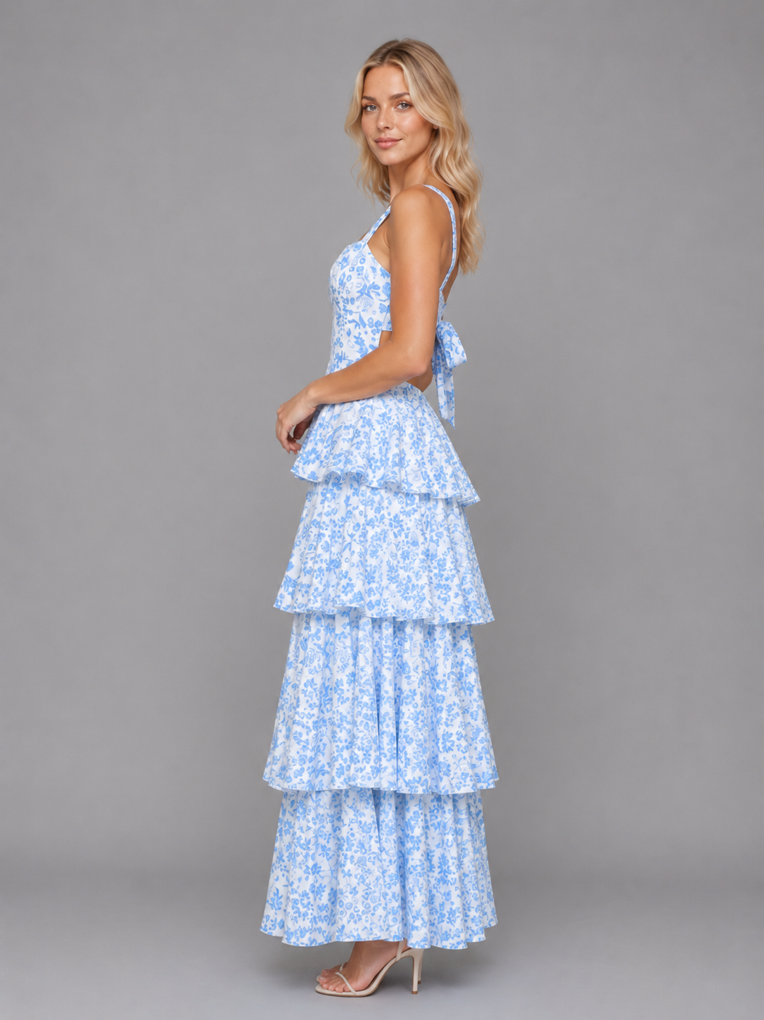 Woman wearing blue and white floral tiered maxi dress with spaghetti straps and layered ruffled skirt, standing against gray background