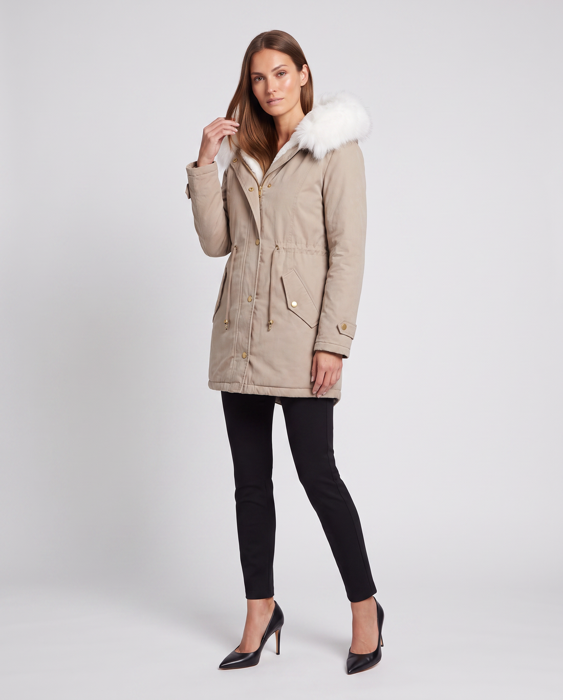 Woman wearing beige winter parka with white fur trim hood, black leggings, and black heels against white background