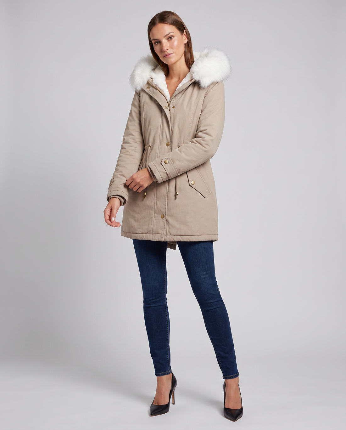 Woman wearing beige hooded winter parka with white fur trim, dark blue jeans, and black heels against neutral background