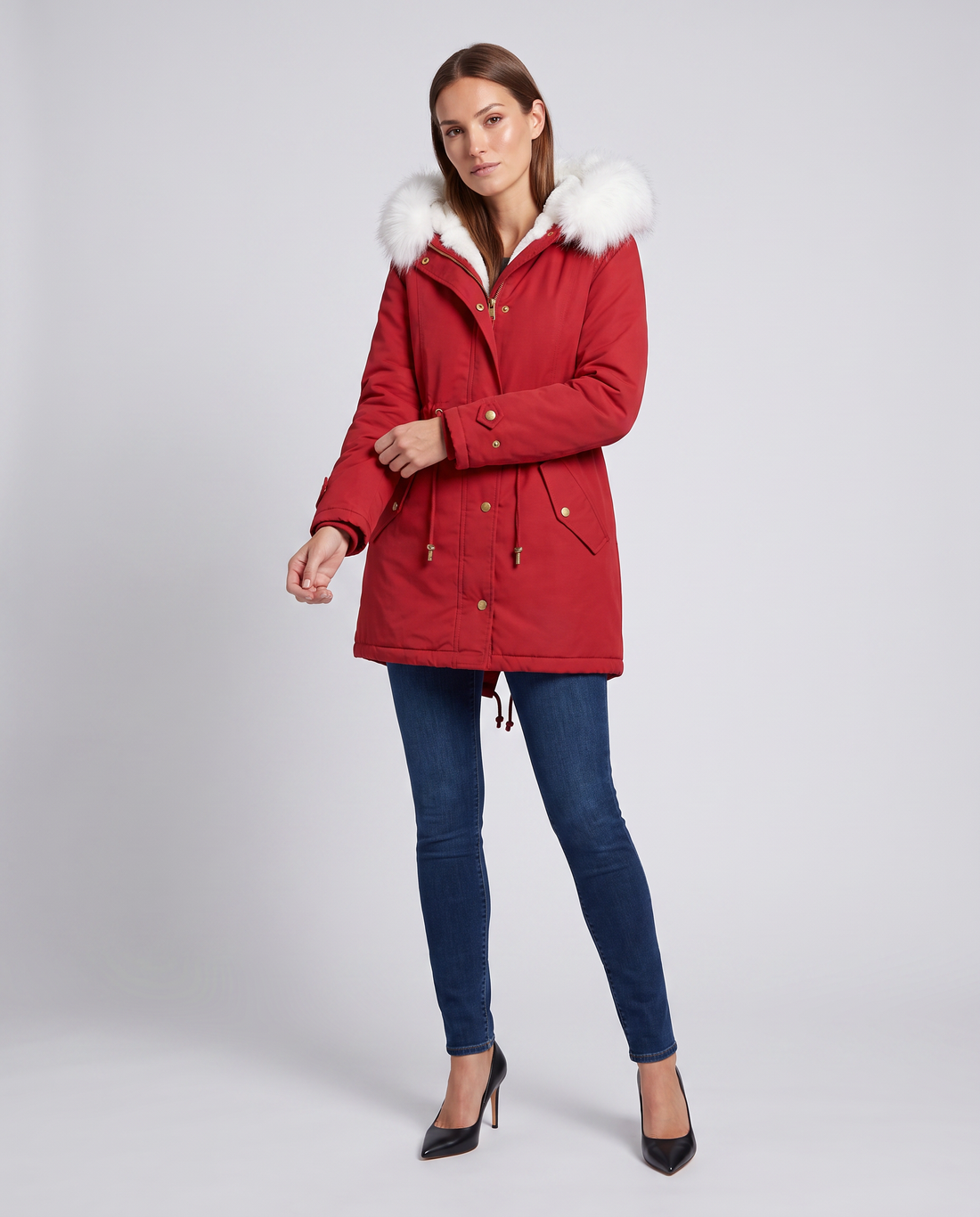 Woman wearing red winter parka with white fur trim hood, dark blue jeans, and black heels