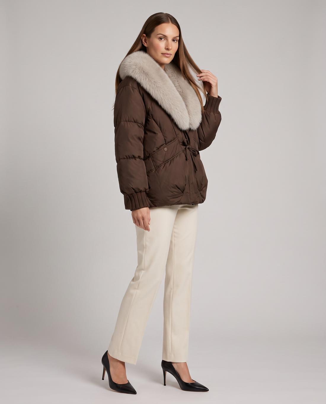 Woman wearing brown puffer jacket with white faux fur collar, cream pants, and black heels
