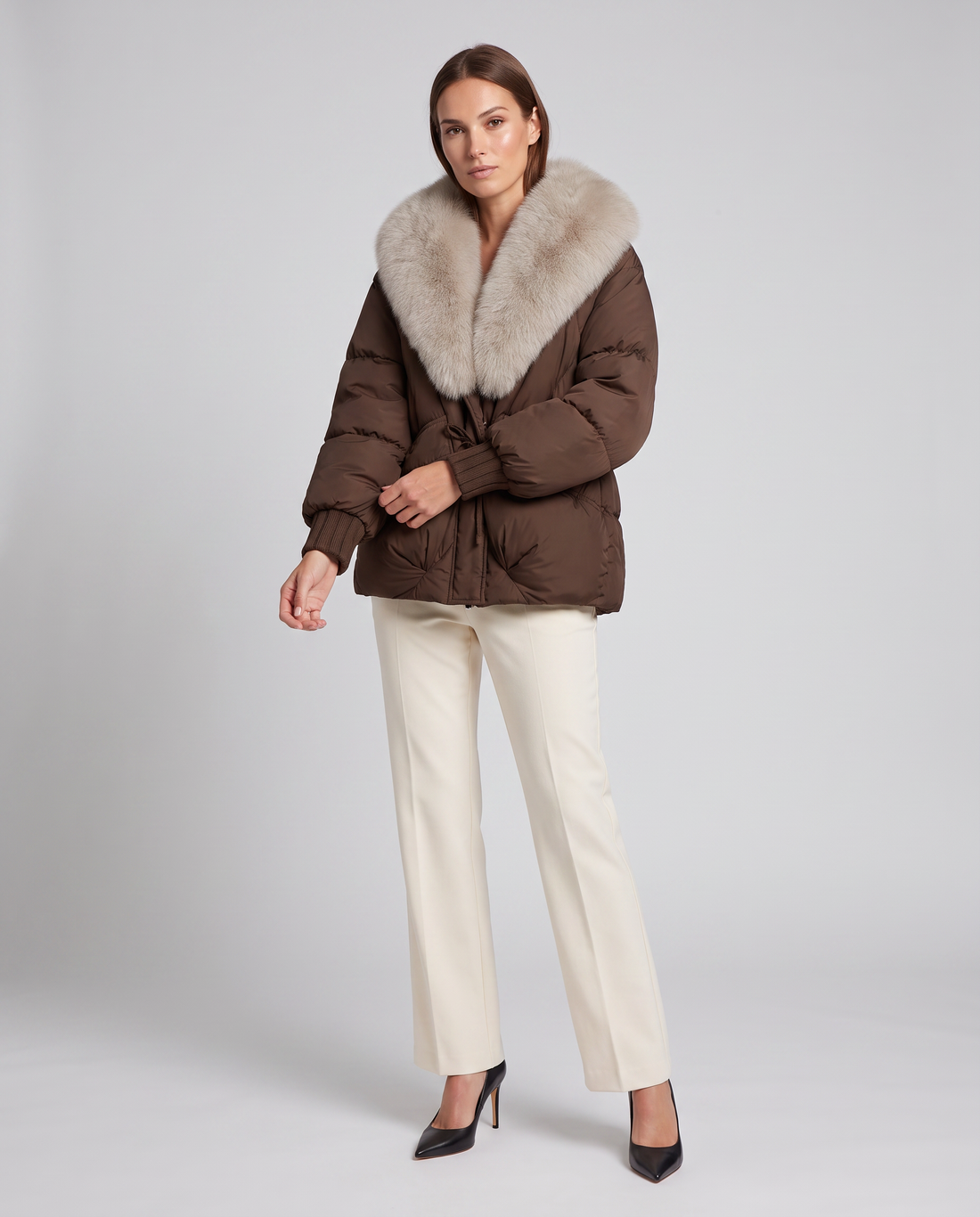 Woman wearing brown puffer jacket with cream faux fur collar and white pants with black heels