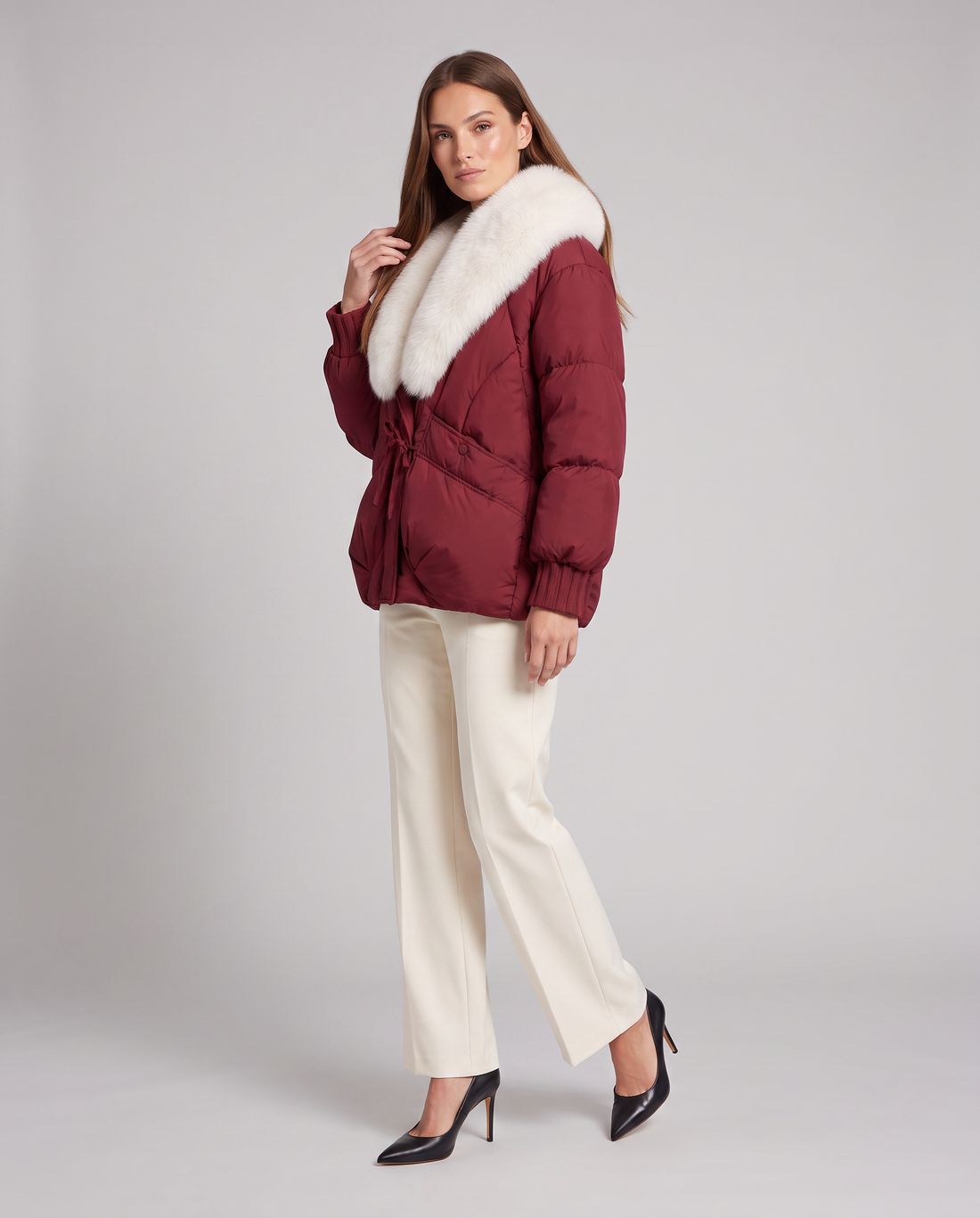 Woman wearing burgundy puffer jacket with white faux fur collar, cream pants, and black heels against gray background